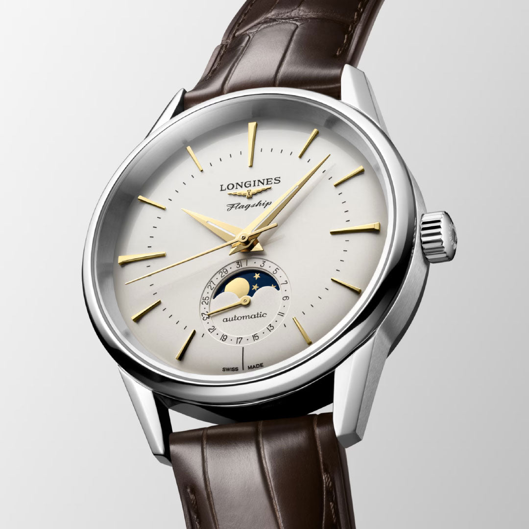 The Longines Flagship Heritage Moonphase Automatic 38.5mm Watch features a white dial, gold hour markers and hands, brown leather strap, and a moon phase display above 6 o’clock—a timeless classic from Longines for any occasion.