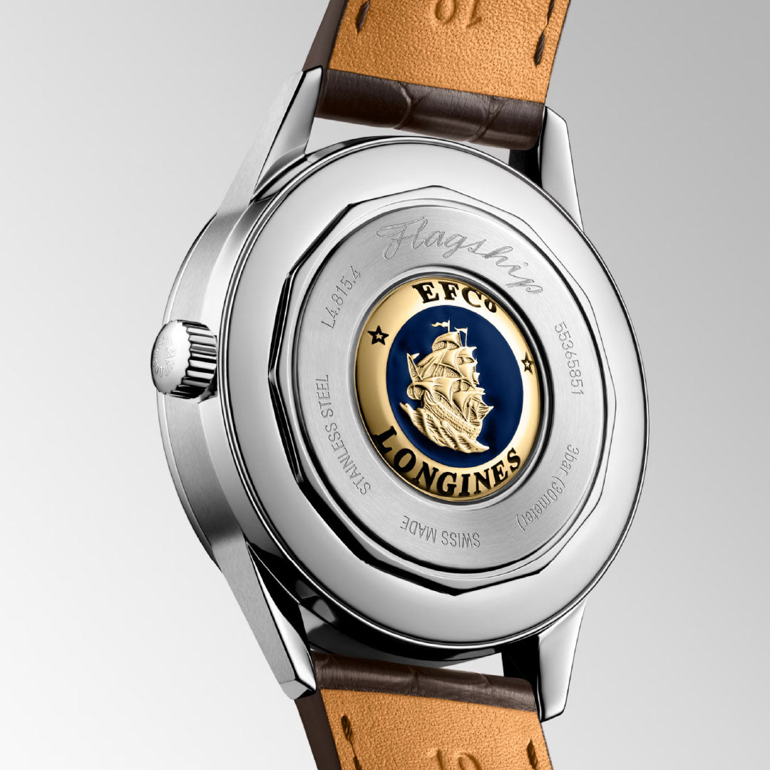 Close-up of the Longines Flagship Heritage Moonphase Automatic 38.5mm, highlighting the engraved gold-and-blue caravel medallion, "Flagship" script, and branding on its stainless steel case with a brown leather strap.