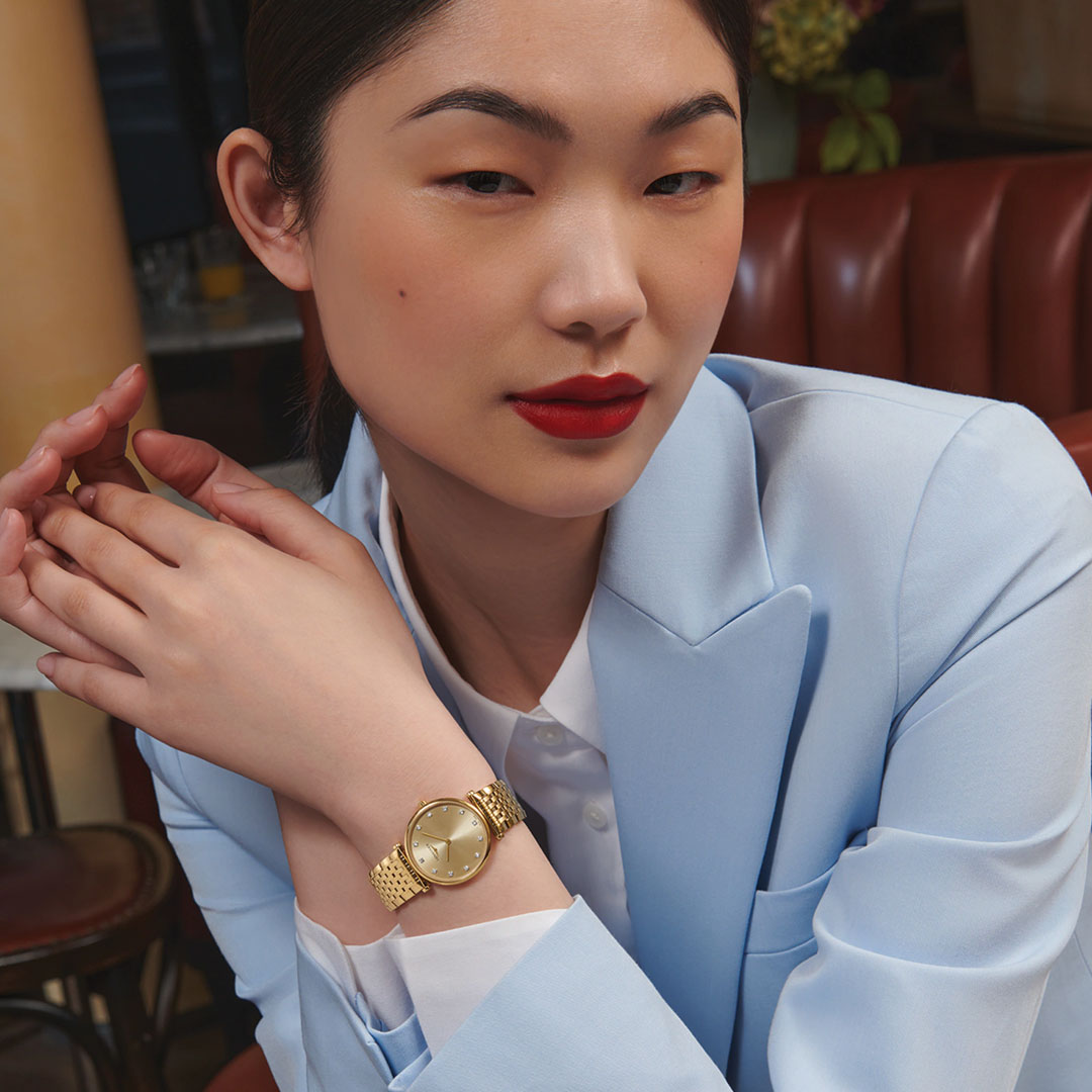 A woman in a light blue blazer and red lipstick poses indoors, showcasing the timeless elegance of the Longines La Grande Classique De Longines Quartz 39mm watch by Longines with its classic gold band on her wrist.