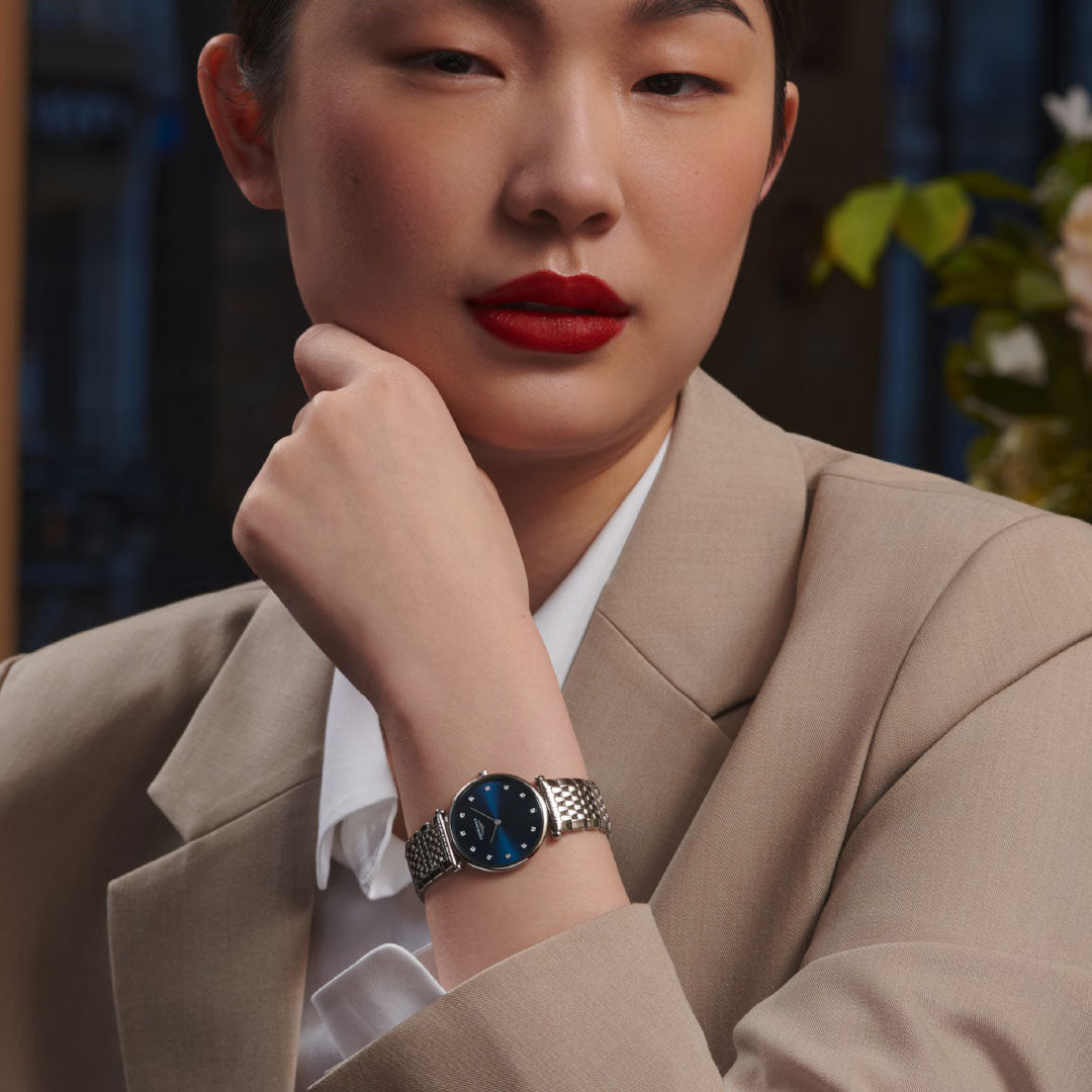 A woman in a beige blazer and white shirt poses with her hand near her face, highlighting the Longines La Grande Classique Quartz 29mm Watch. Her red lipstick and confident look add to the classic elegance of this Longines timepiece.