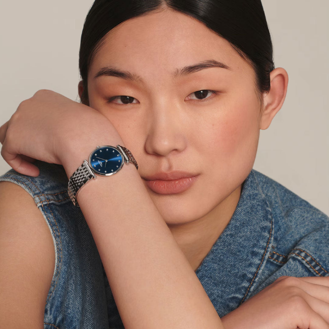 A person with straight black hair in a sleeveless denim jacket rests their head on their arm, highlighting the elegant Longines La Grande Classique Quartz 29mm Watch with its dark blue face against a neutral background.