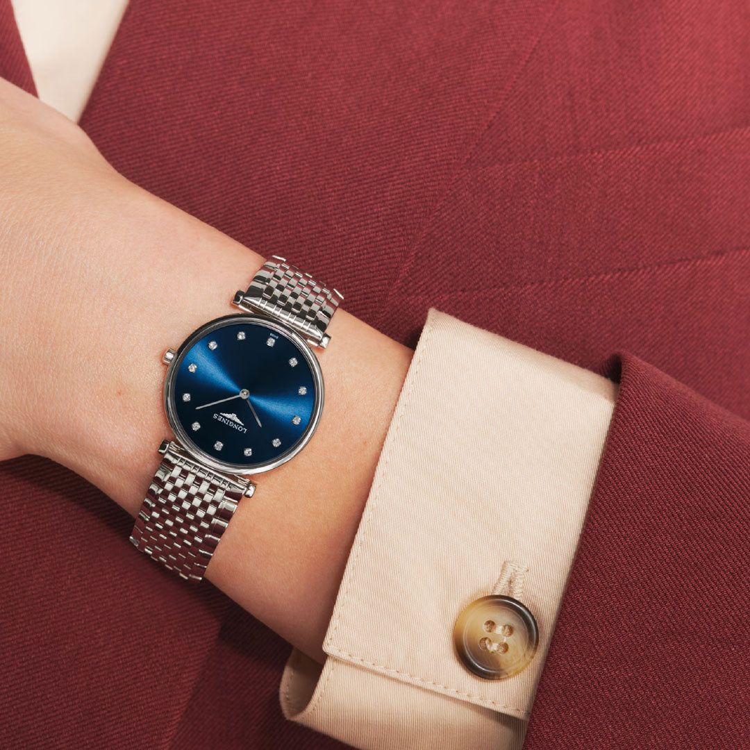 A person in a maroon blazer and cream shirt wears a Longines La Grande Classique Quartz 29mm watch with a blue dial and metal band on their left wrist.