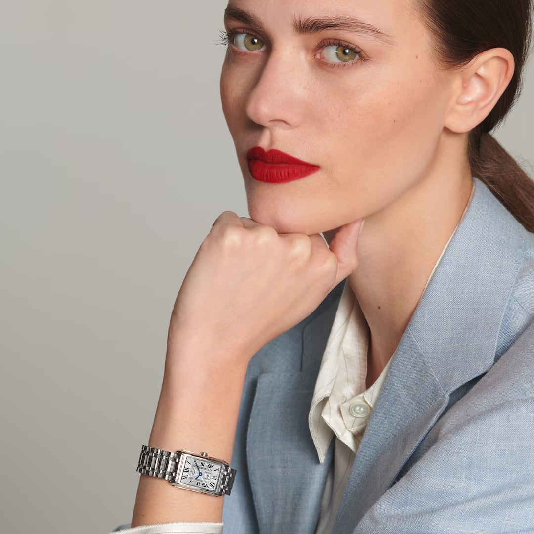 Sporting a light blue blazer and white shirt, a person gazes thoughtfully while resting their chin on their hand. The Longines Dolcevita Quartz 20.80 X 32mm Watch with its rectangular case exudes timeless elegance, complemented by bright red lipstick against the neutral background.