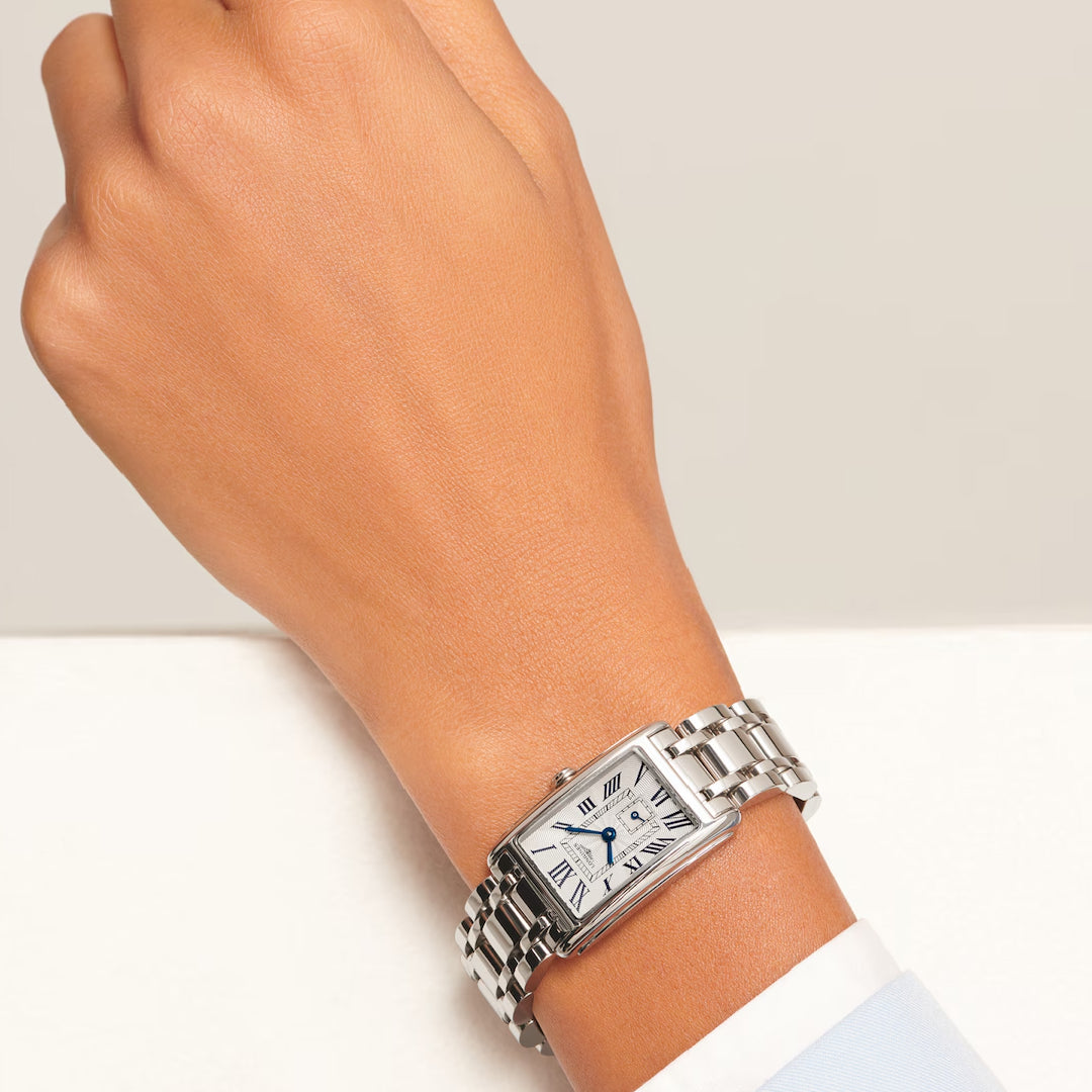 A hand adorned with the Longines Dolcevita Quartz 20.80 X 32mm watch, featuring a sleek silver rectangular case, white dial with Roman numerals, and a metal link bracelet, pairs elegantly with the cuff of a light blue shirt.