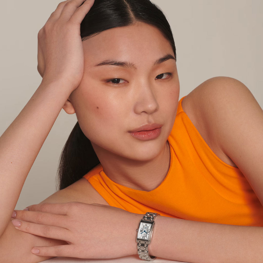 A woman with straight black hair pulled back rests her head on one hand, crossing her arms, and wears a sleeveless orange top. She showcases the timeless elegance of the Longines Dolcevita Quartz 20.8 x 32mm watch by Longines.