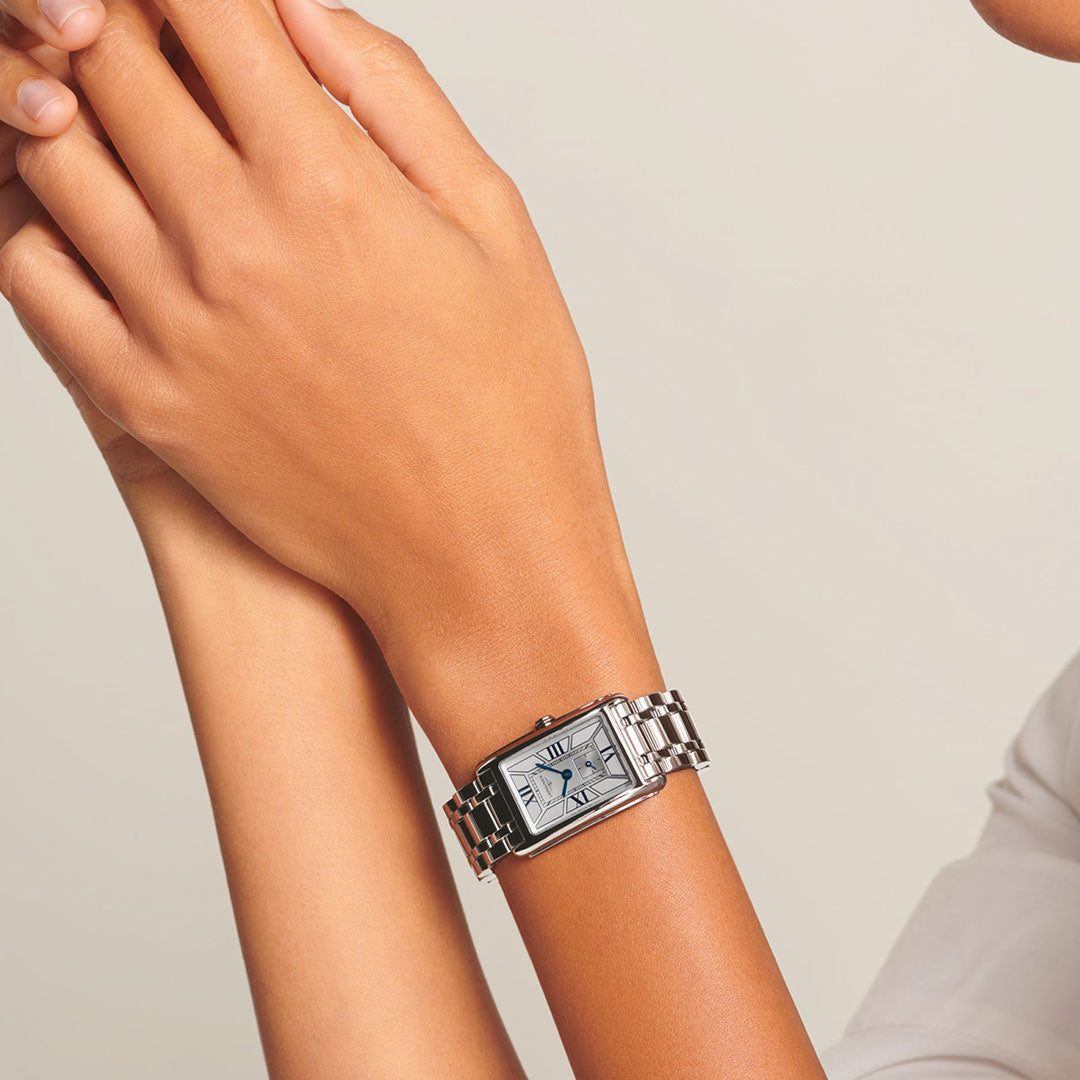 A person with crossed arms wears a Longines Dolcevita Quartz 20.8 x 32mm Watch, a silver rectangular timepiece by Longines with a white dial and Roman numerals, shown against a plain light background for timeless elegance.