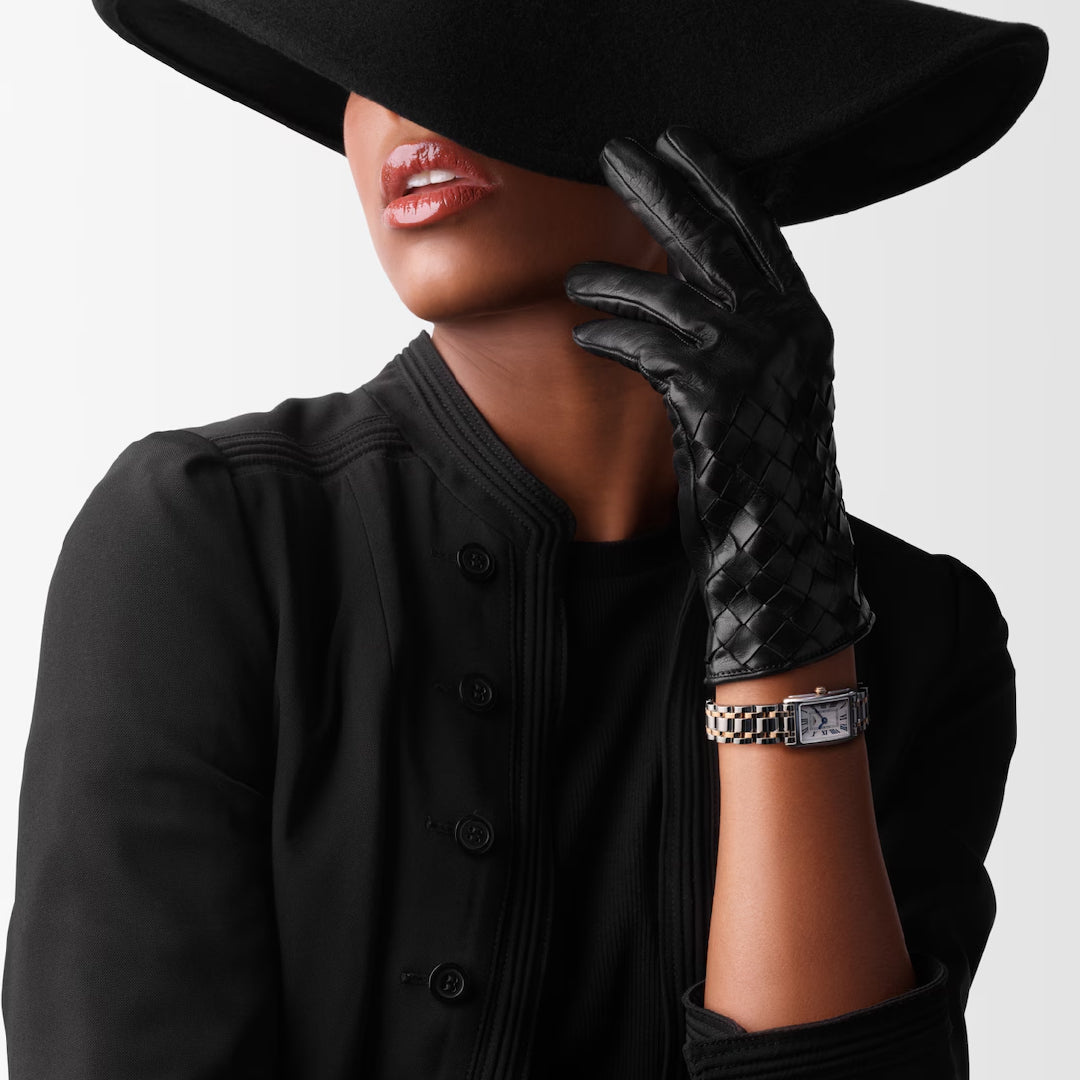 A person in a black wide-brimmed hat and gloves raises an arm, flaunting the timeless elegance of the Longines Dolcevita Quartz 17.70 X 27mm watch. The rectangular case shines brightly, perfectly complementing their stylish black jacket for a sophisticated appearance.