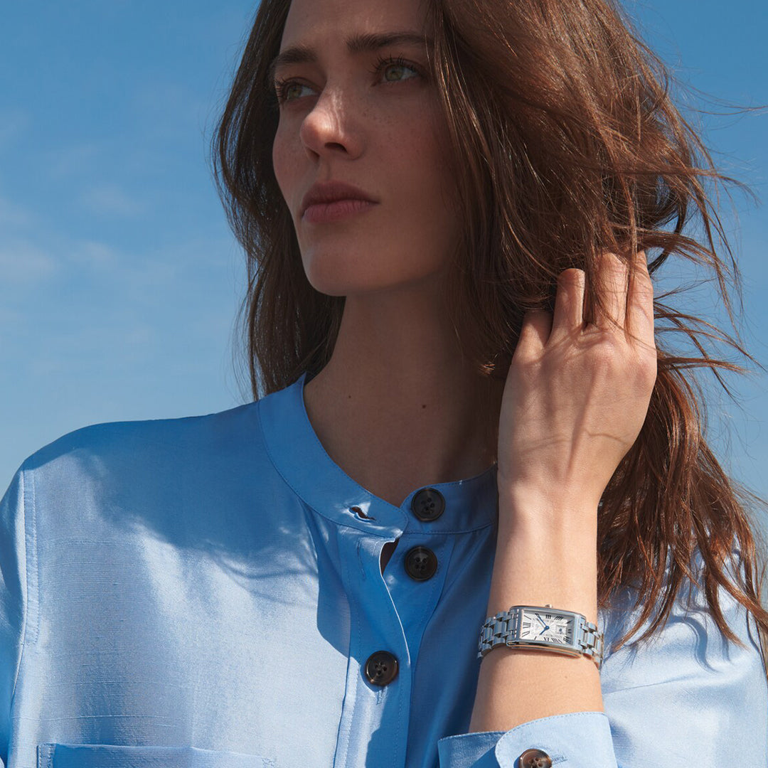 A woman with long brown hair wears a light blue blouse with a high collar and wooden buttons, gazing to the side against a clear blue sky. On her wrist is a Longines Dolcevita Quartz 23.30 X 37mm Watch, featuring a stylish silver band and rectangular case, exuding timeless elegance.