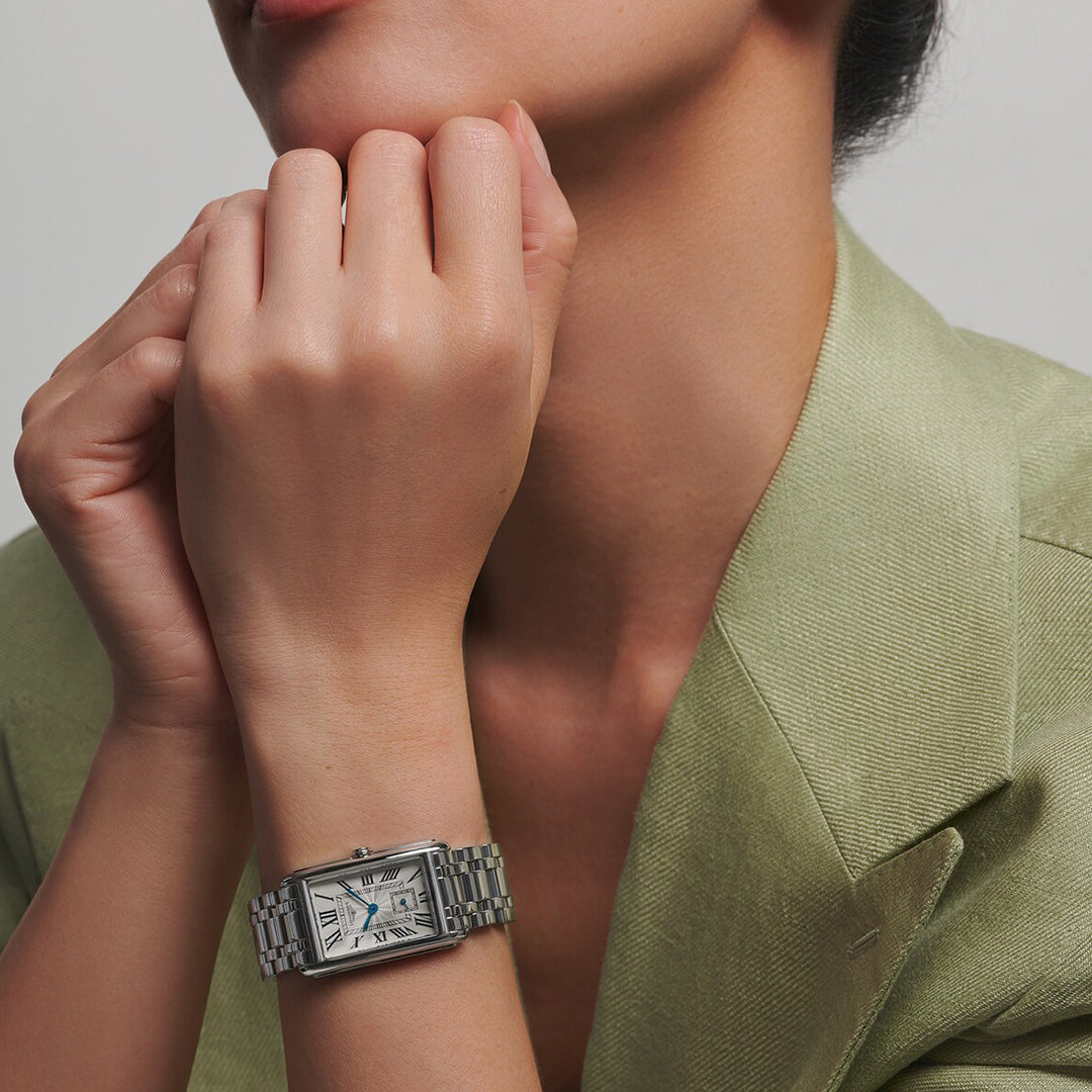 A person in a light green blazer rests their chin on their hands, exemplifying the timeless elegance of the Longines Dolcevita Quartz 23.30 X 37mm watch. It features a silver rectangular case and a white dial with Roman numerals, set against a plain background.