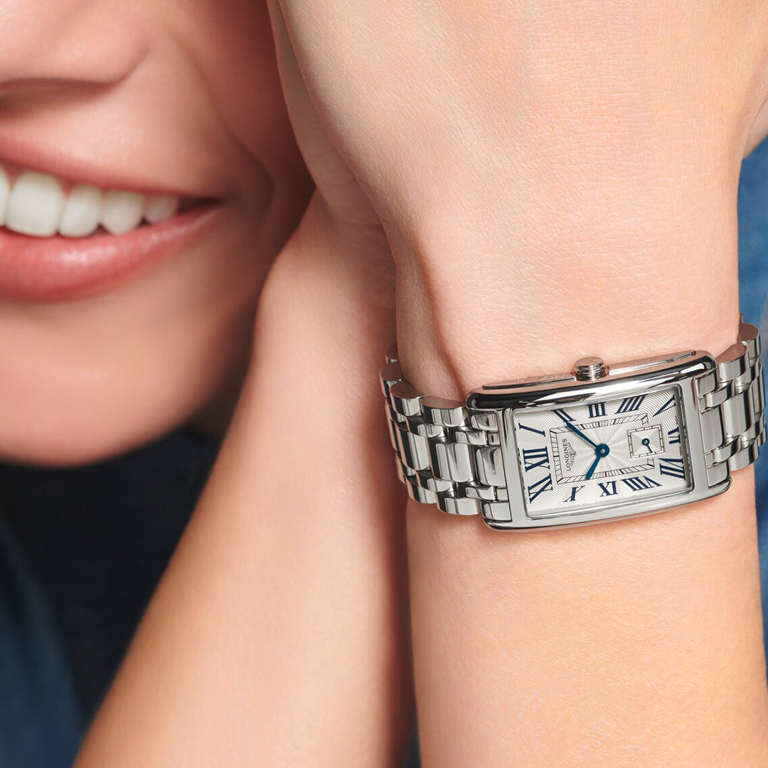 A person exudes timeless elegance, smiling gently with their chin on their hand. They wear a Longines Dolcevita Quartz 23.30 X 37mm watch, featuring a rectangular case with Roman numerals and a metal link band. Their blue top complements the watch perfectly.