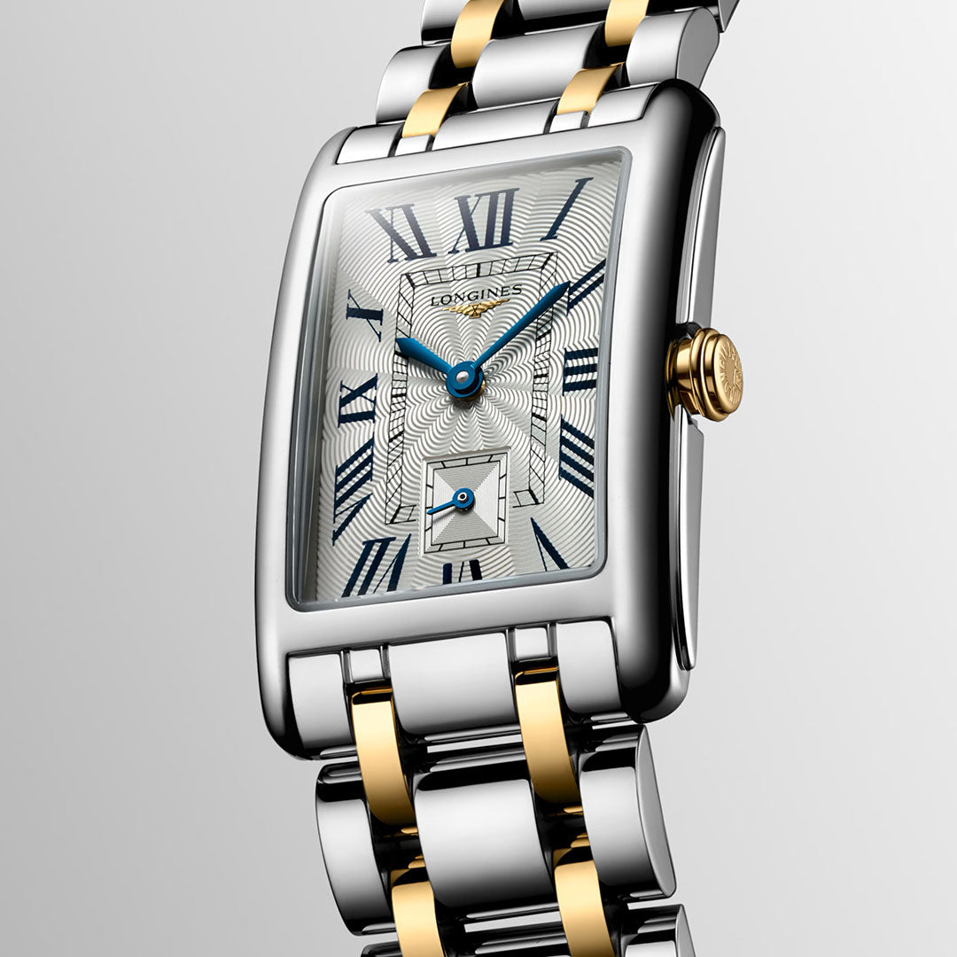 A close-up of the Longines Dolcevita Quartz 23.3 x 37mm Watch shows its rectangular silver face with blue Roman numerals, gold accents, and a two-tone metal band, highlighting Longines' timeless elegance against a light background.