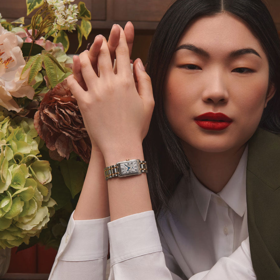 A woman with long dark hair and bold red lipstick poses beside a vase of hydrangeas, wearing a white blouse, green blazer, and the Longines Dolcevita Quartz 20.8 x 32mm Watch on her wrist.