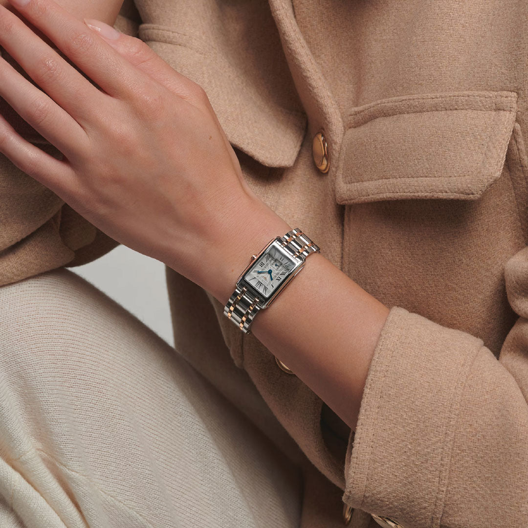 A person in a beige coat and cream pants rests their hand on their knee, showcasing the Longines Dolcevita Quartz 20.8 x 32mm watch with its signature rectangular case and metal bracelet by Longines.