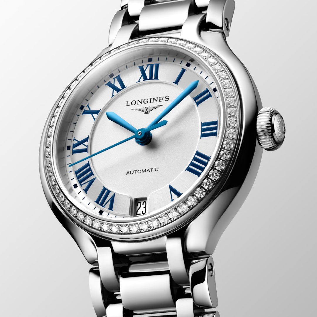 A close-up of the Longines Primaluna Automatic 34mm watch featuring a silver band, blue Roman numerals and hands, a diamond-set bezel, and a date window at 6 o'clock, set against a light gray background.