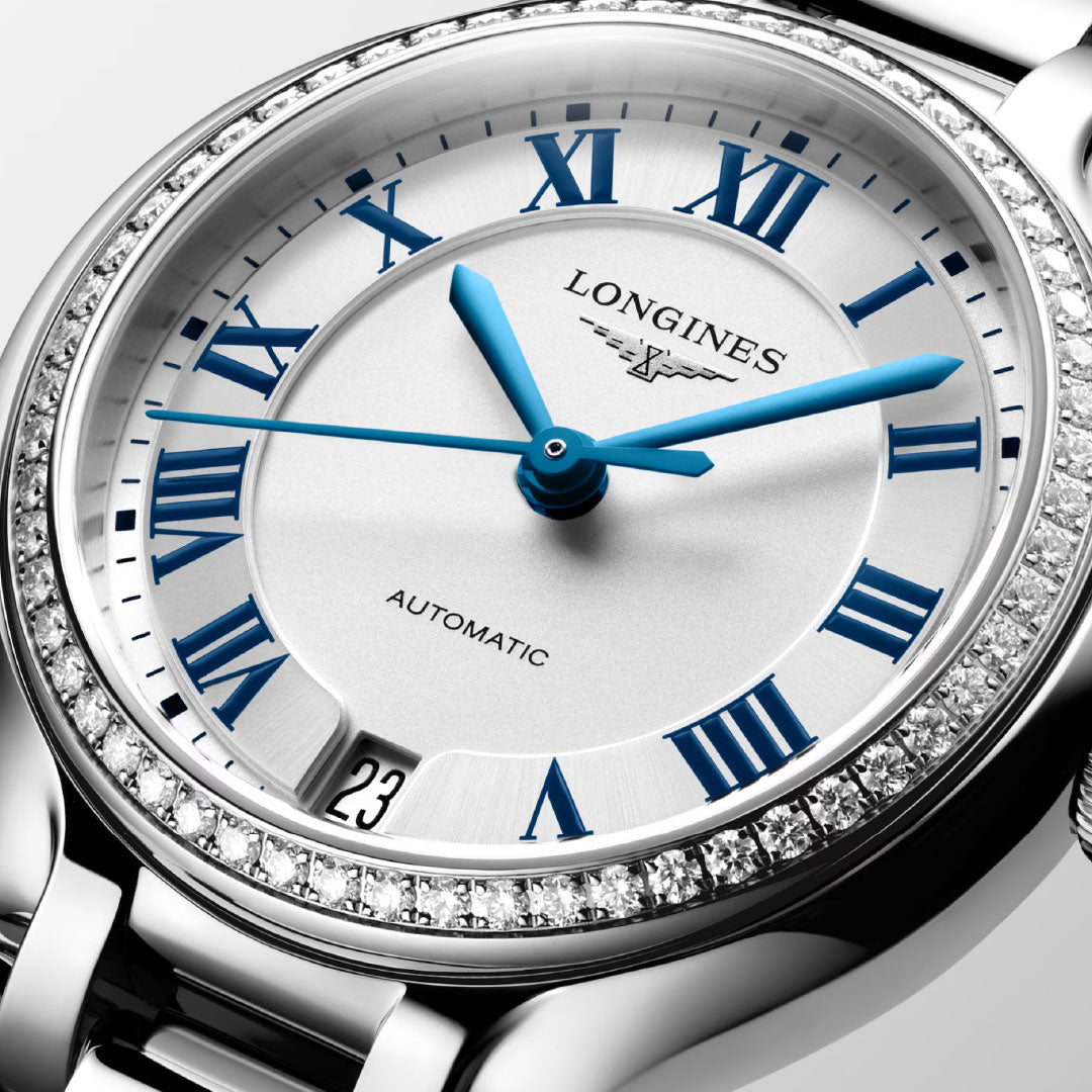 Close-up of a Longines Primaluna Automatic 34mm women’s watch featuring a diamond-set bezel, blue Roman numeral markers and hands, and a date window at 3 o’clock. The silver dial is labeled "AUTOMATIC" for added elegance.