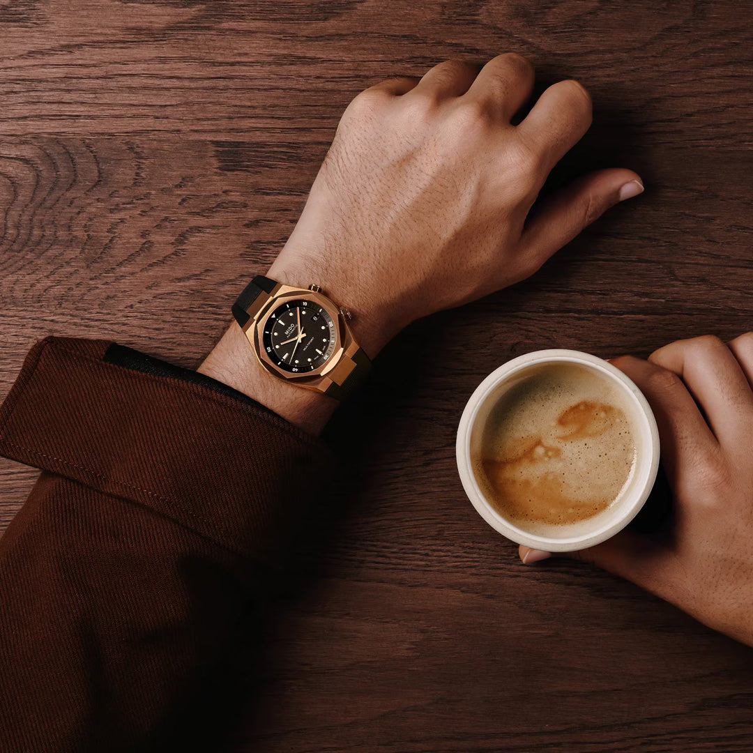 A person in a brown jacket wears a MIDO Multifort 8 Two Crowns Automatic 40mm Watch on their left wrist and holds a cup of coffee with their right hand, all set on a wooden surface.