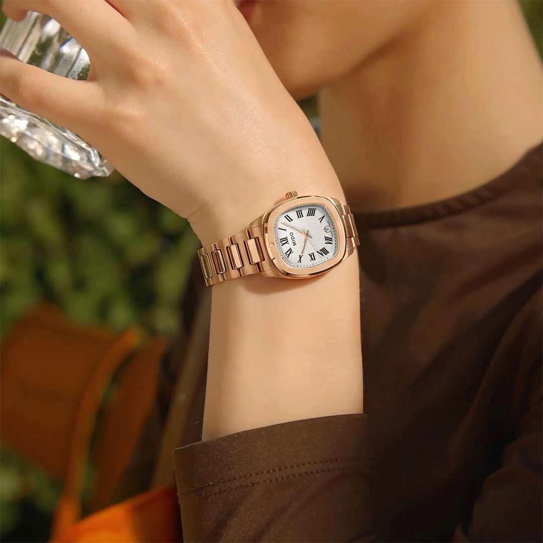 A person wearing a MIDO Multifort TV Quartz 28mm Watch with a gold case, white face, and Roman numerals holds a clear glass of water against a backdrop of blurred greenery.