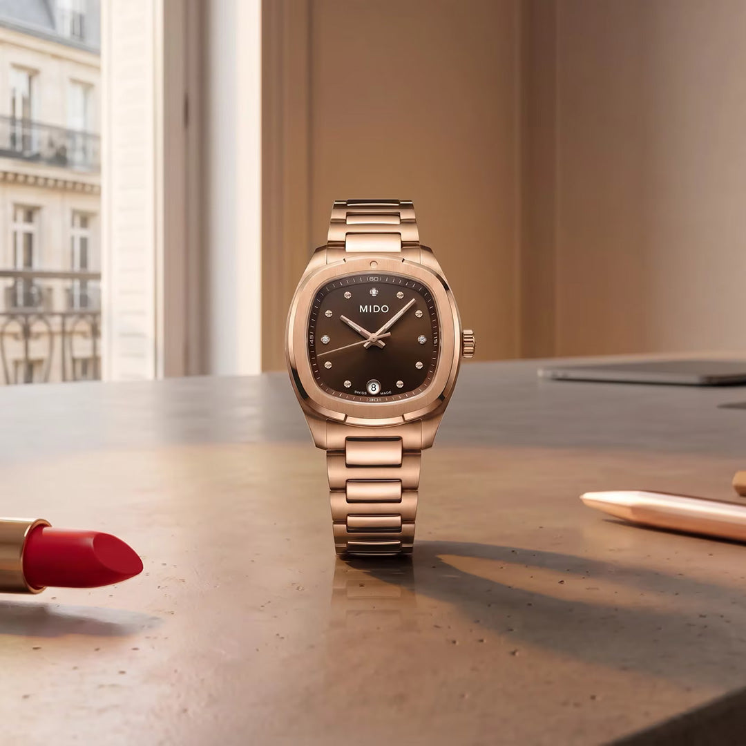 A MIDO Multifort TV Quartz 28mm Watch in rose gold with a brown dial rests on a desk beside a red lipstick and a white pen. In the background, a window reveals part of a building.