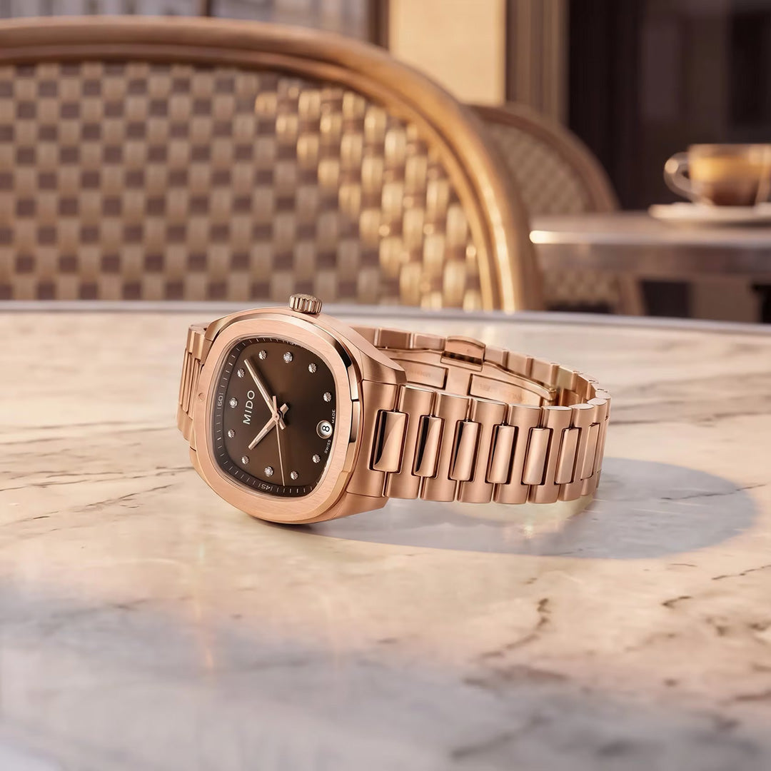 A MIDO Multifort TV Quartz 28mm watch in rose gold with a black dial rests on a marble table at an outdoor café, framed by woven chairs and a cup of coffee in the softly blurred background, showcasing the timeless elegance of the MIDO brand.