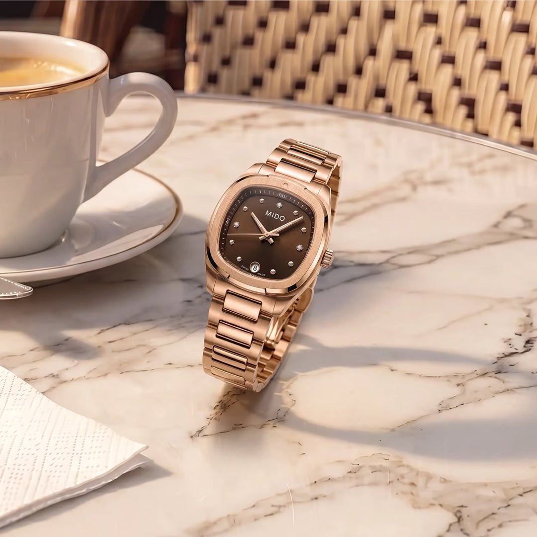 A MIDO Multifort TV Quartz 28mm watch in rose gold rests on a marble café table beside a cup of coffee, a wicker chair, and a white napkin, with sunlight casting soft shadows.