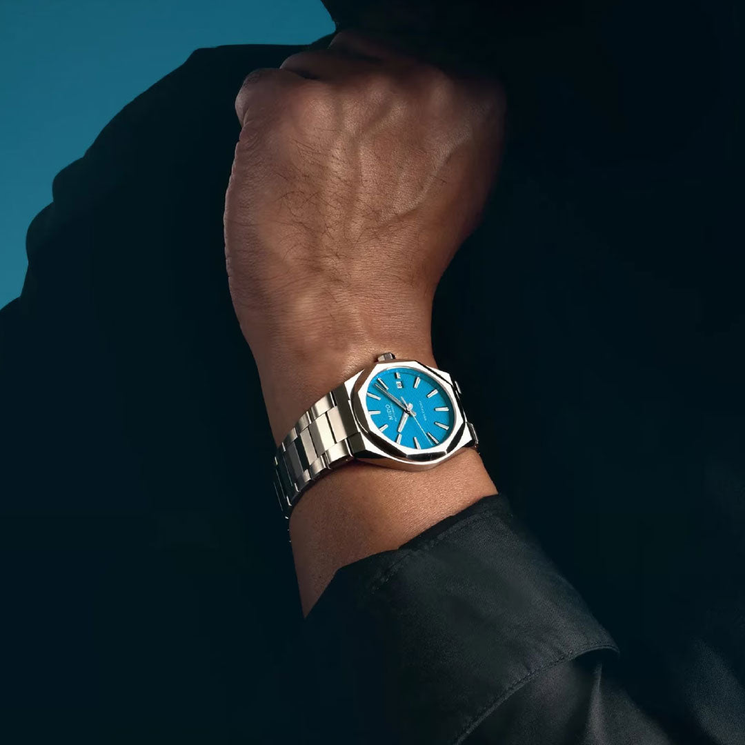 A person wears the MIDO Multifort 8 One Crown Automatic 40mm Watch, featuring a silver octagonal bezel and vibrant blue face. Their bent arm is clad in a dark long-sleeve shirt, set against a deep blue background.