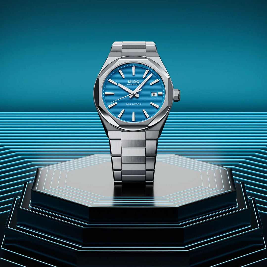 The MIDO Multifort 8 One Crown Automatic 40mm Watch stands upright on an illuminated black platform with blue neon accents, highlighting its silver case, octagonal bezel, and blue face against a gradient blue background.