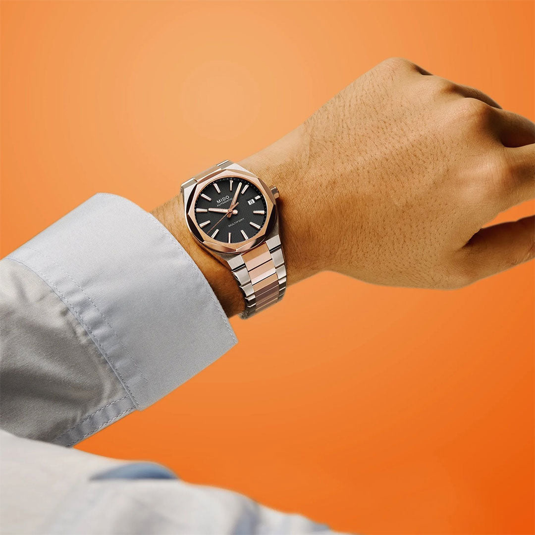 A person wears a MIDO Multifort 8 One Crown Automatic 40mm watch with a silver case, black dial, and sapphire crystal on their left wrist, paired with a light blue shirt against an orange background.
