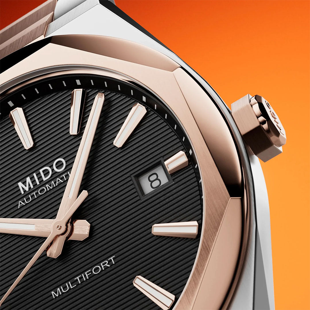 Close-up of the MIDO Multifort 8 One Crown Automatic 40mm Watch, featuring a black textured dial, rose gold hands and markers, and a date window showing 8, set against an orange gradient background.