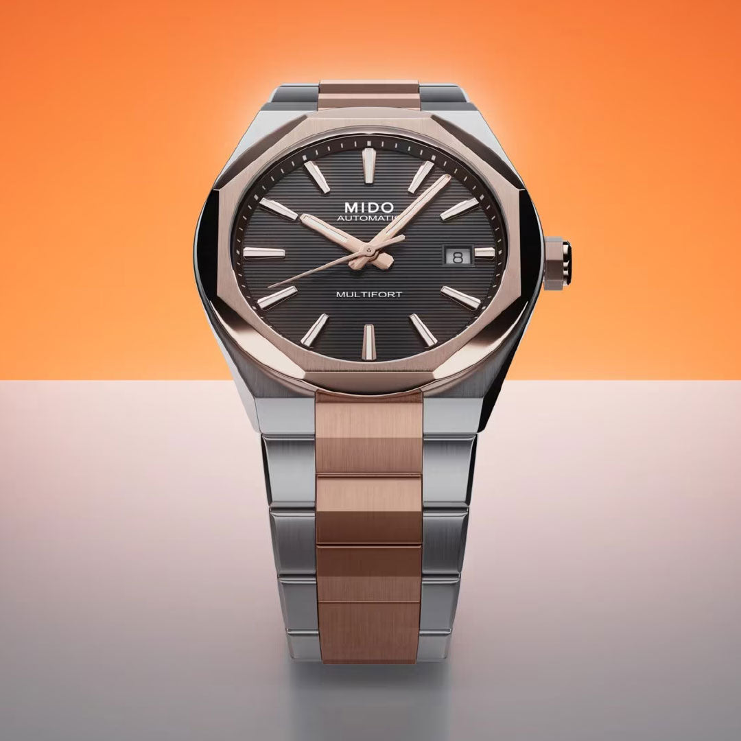 The MIDO Multifort 8 One Crown Automatic 40mm Watch features a silver and rose gold metal band, octagonal bezel, black textured dial with date window and silver hour markers, displayed on a gradient orange background.