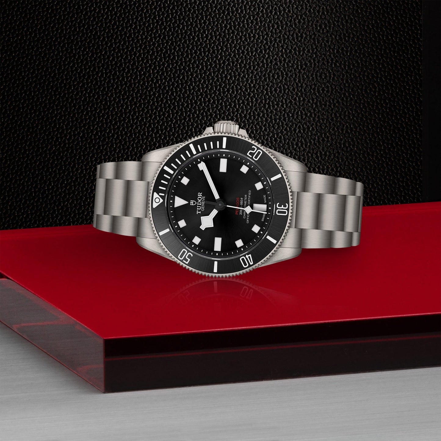 The TUDOR Pelagos 39 is a sleek titanium wristwatch featuring a black dial with large hour markers and a date window at the 3 o'clock position. Its stainless steel band complements the red and black textured backdrop, making it an ideal sports watch for those who value style and precision.