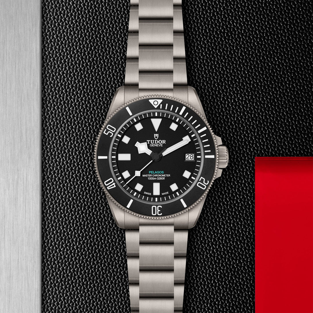 The TUDOR Pelagos Ultra, a silver wristwatch with a black face and luminescent markers set to 10:11, features a date function. It rests on a textured black background with a red accent on the right, bearing "TUDOR" at the top.