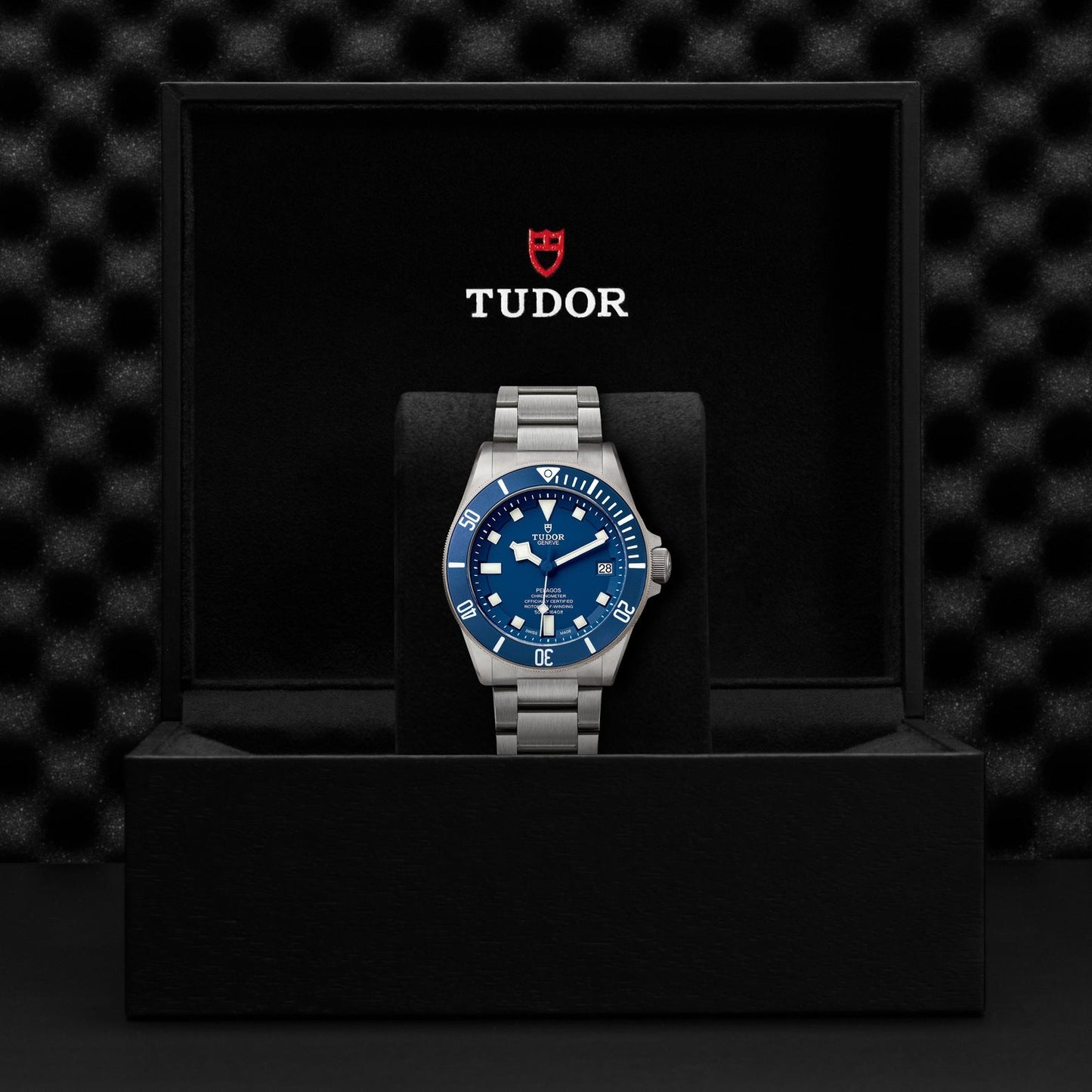 The TUDOR Pelagos, a luxury divers' watch featuring a blue dial and a titanium bracelet, is elegantly displayed in an open black box adorned with the TUDOR logo on the lid. A textured black background enhances its sophisticated presentation, emphasizing the watch's waterproof design.