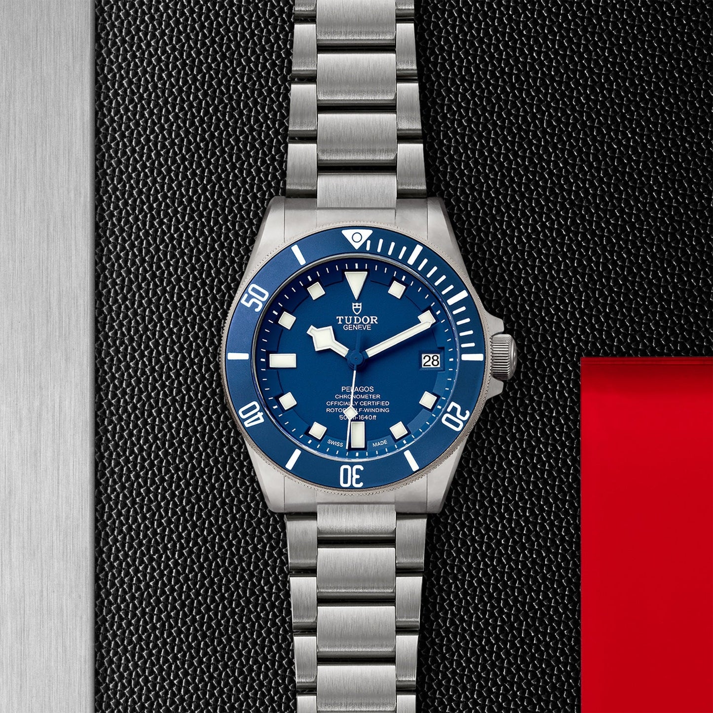 Close-up of the TUDOR Pelagos, a stainless steel divers' watch featuring a blue dial and bezel with white hour markers and a date display at 3 o'clock. It rests on a textured black and silver surface, accented with red on the right, enhancing its sophisticated design.