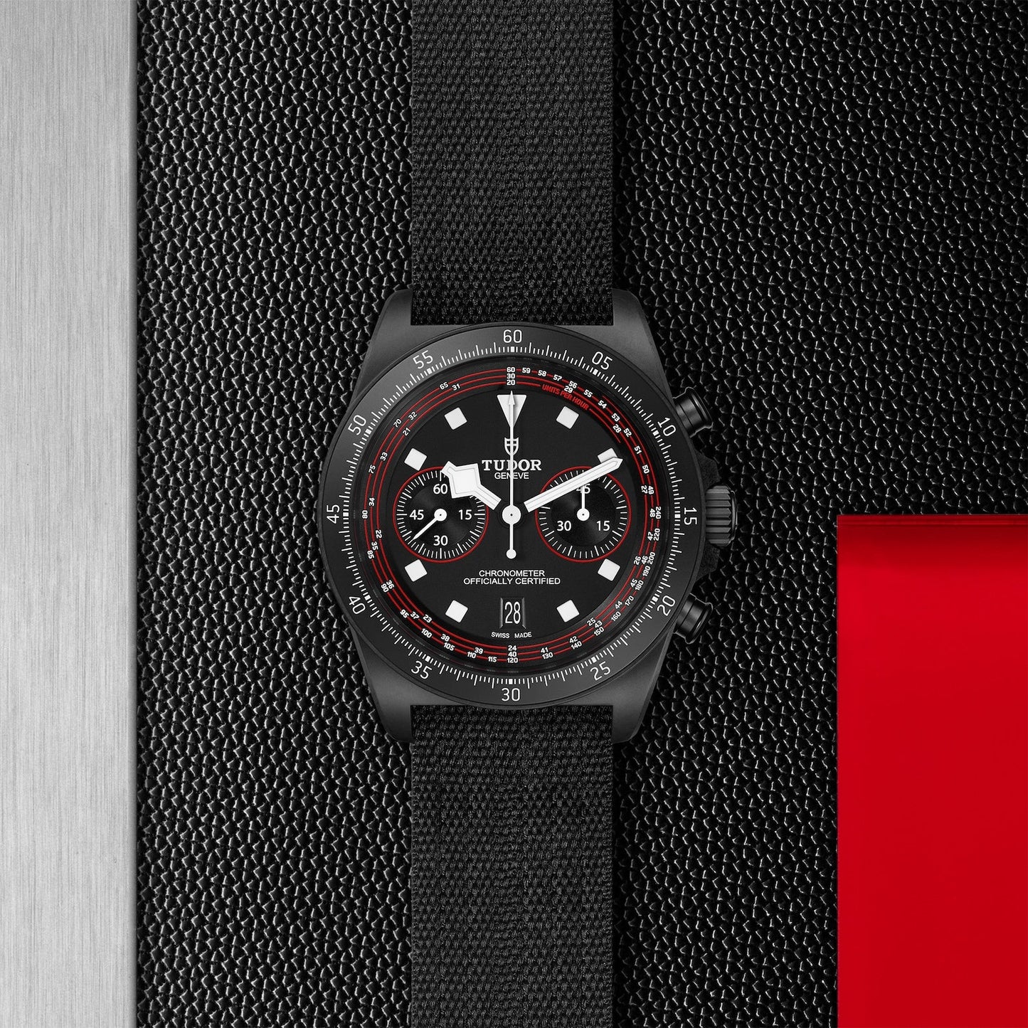 The TUDOR Pelagos FXD Chrono, by TUDOR, is a black timepiece featuring a cycling-themed chronograph and textured strap. It includes three sub-dials and showcases a carbon composite case with red and white accents on a textured black and metallic background, complemented by a distinctive red square accent.