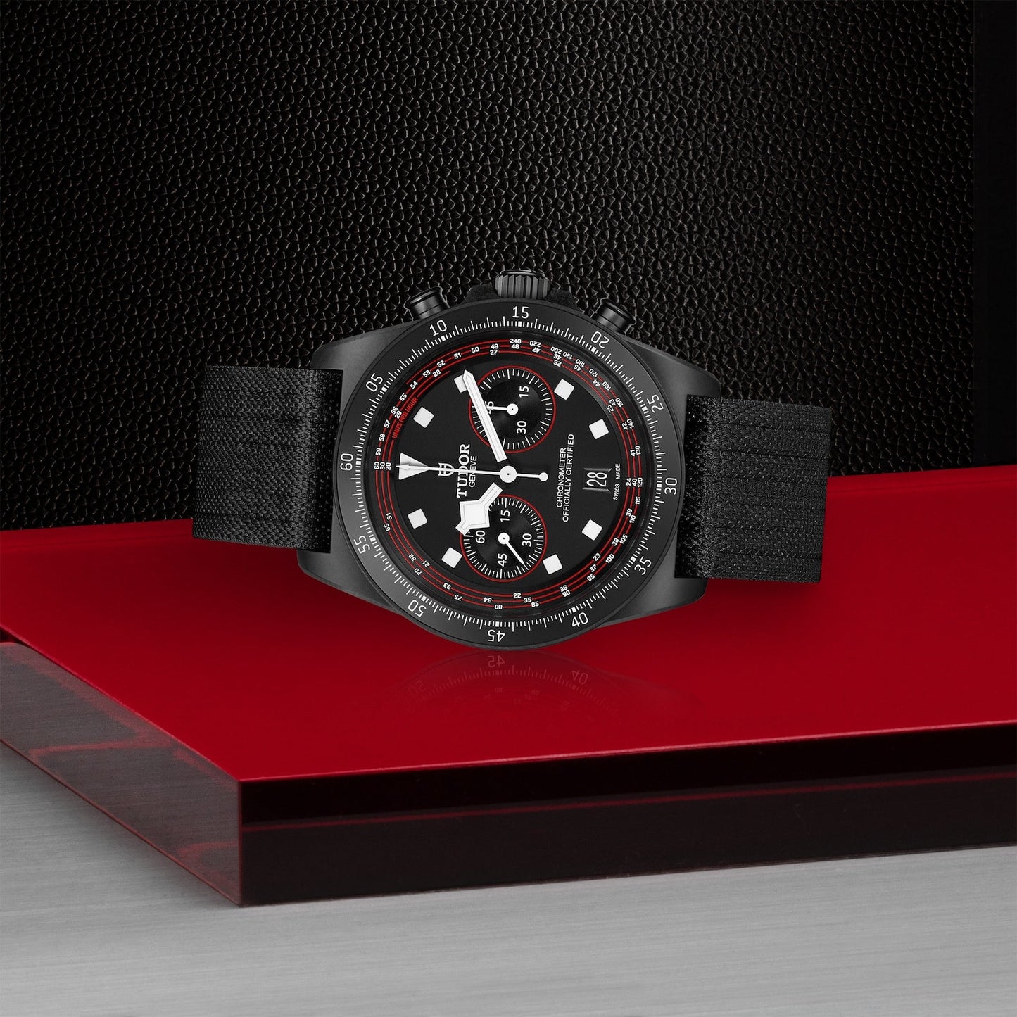 A TUDOR Pelagos FXD Chrono wristwatch, featuring a black fabric strap and a dial with red and white cycling-themed chronograph and date display, is showcased on a sleek red and black surface against a textured black background.