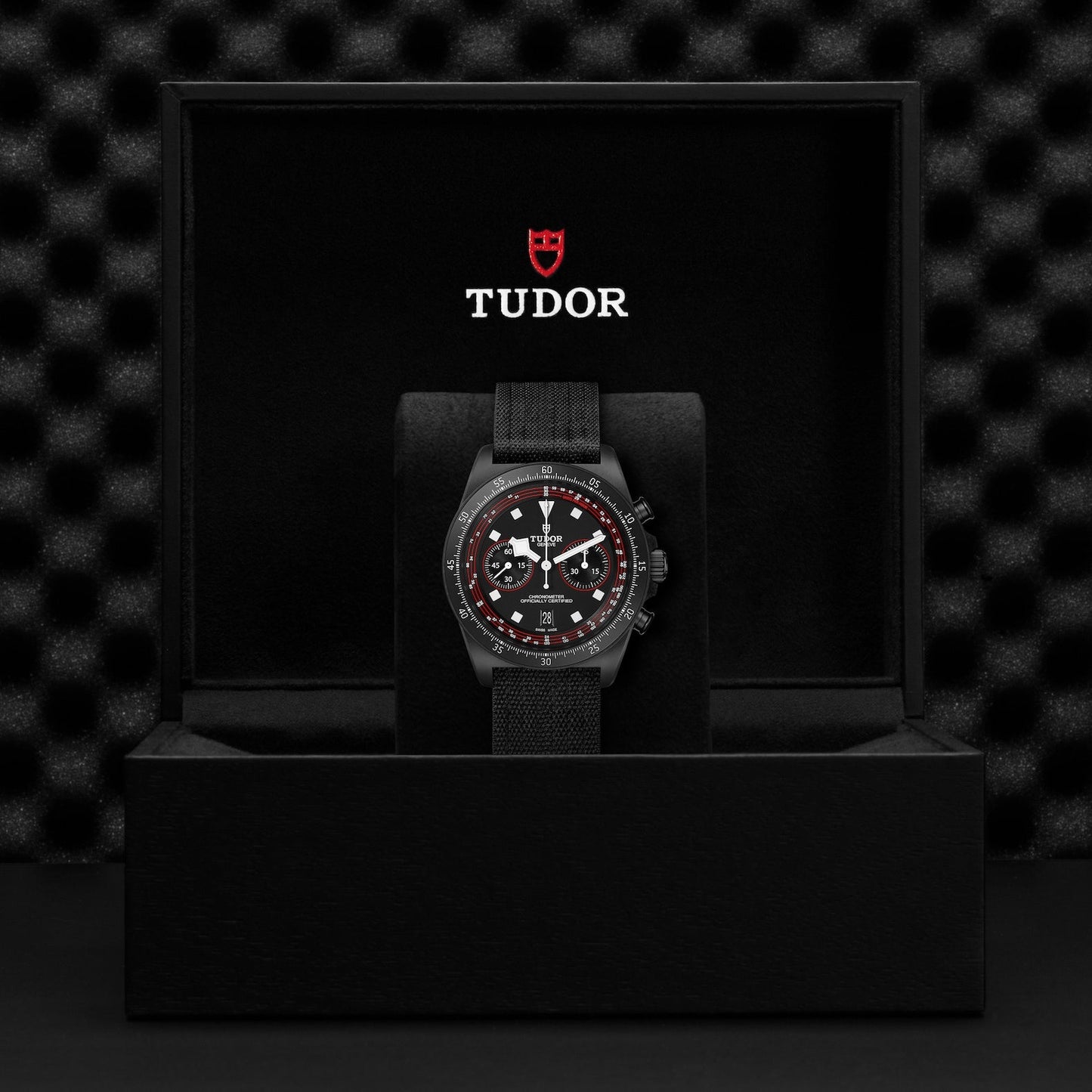 A TUDOR Pelagos FXD Chrono watch in black, featuring a red and white detailed dial, is displayed on a black cushion inside an open black box. The interior of the box lid showcases the TUDOR logo in red and white. The backdrop has a textured black finish that complements the cycling-themed chronograph design.