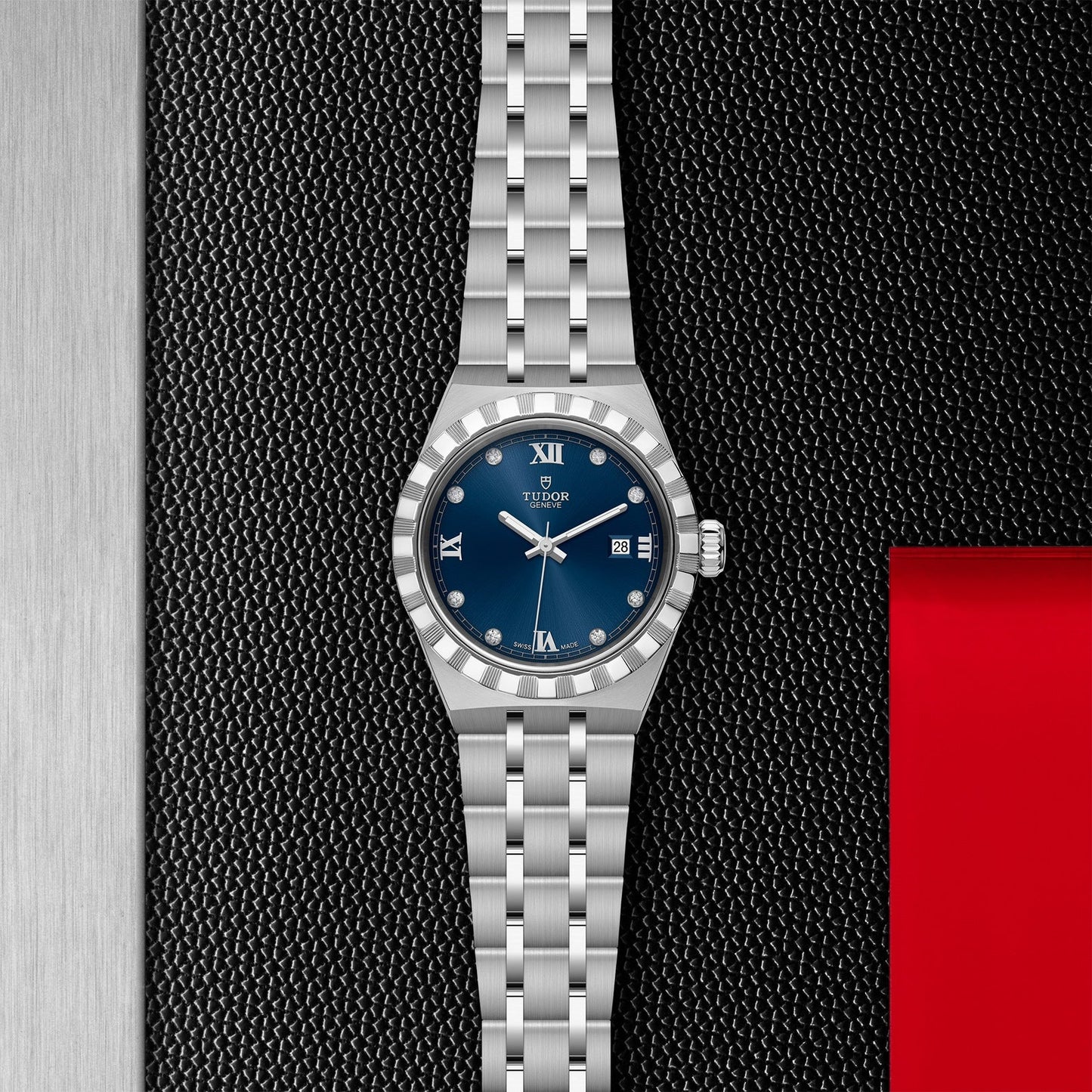 Set against a textured black background, the TUDOR Royal from TUDOR showcases an automatic sport-chic design featuring a metal bracelet and blue dial. Its elegance is highlighted by Roman numerals and a patterned silver bezel, while the red rectangle provides striking contrast.