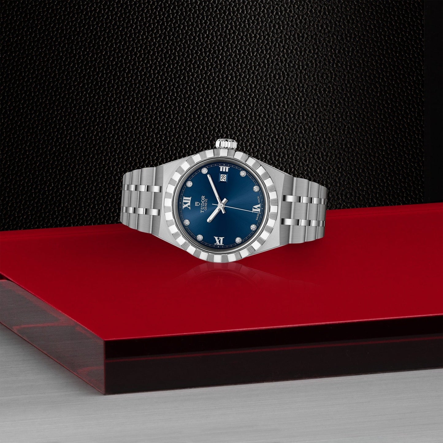 The TUDOR Royal wristwatch by TUDOR, featuring a silver case, blue dial with Roman numerals, and a date display, rests elegantly on a red surface against a textured black backdrop. This automatic sport-chic design beautifully demonstrates the seamless fusion of style and functionality.