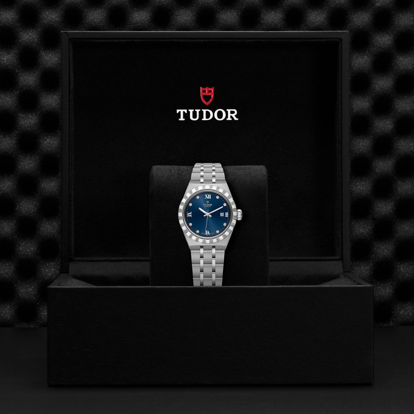 Inside an open black box, a TUDOR Royal wristwatch catches the eye with its stunning blue dial and date display. Crafted from stainless steel and gold, it exudes sport-chic elegance. The interior of the box is lined with black fabric, featuring a textured black backdrop that showcases the red TUDOR logo on the inside lid.