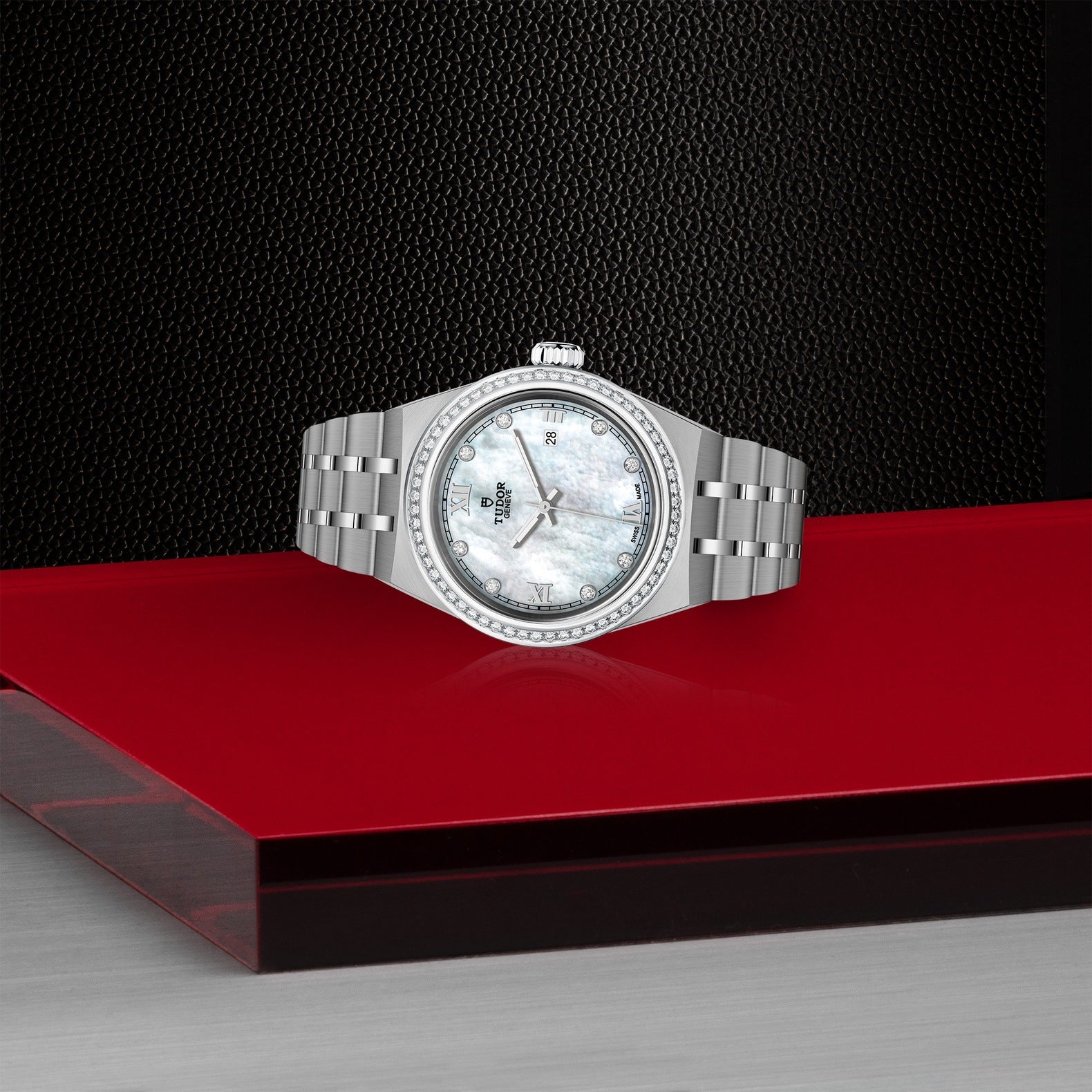 A TUDOR Royal wristwatch in silver, featuring a mother-of-pearl face and diamond bezel, is displayed on a red and black platform. This automatic sport-chic timepiece boasts a stainless steel band and Roman numerals against a textured black background.