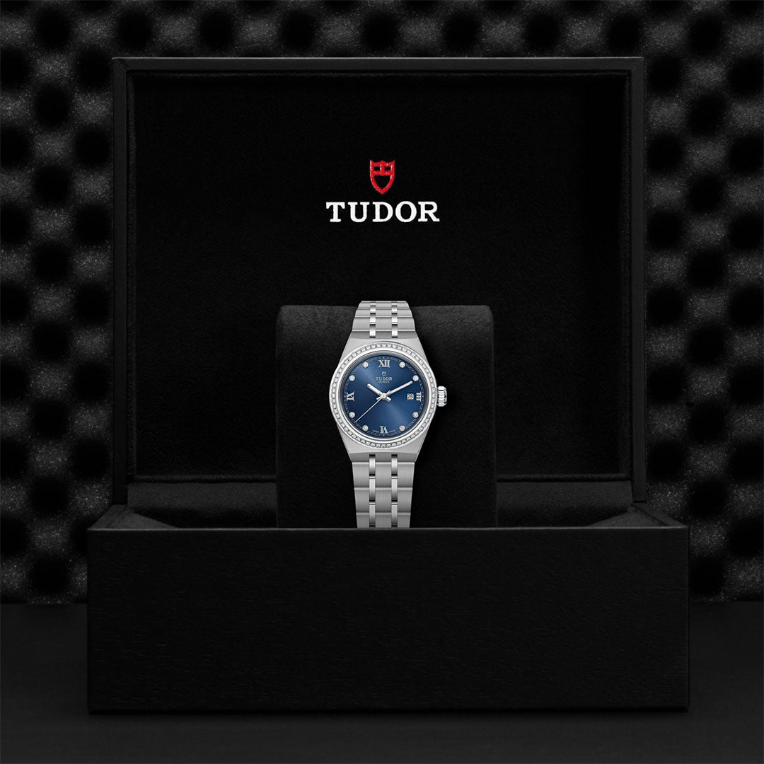 The TUDOR Royal, an automatic watch by TUDOR with a blue dial and silver bracelet, is showcased in an open black box featuring the Tudor logo and red shield on the lid, set against a dark, textured background to emphasize its sport-chic design.