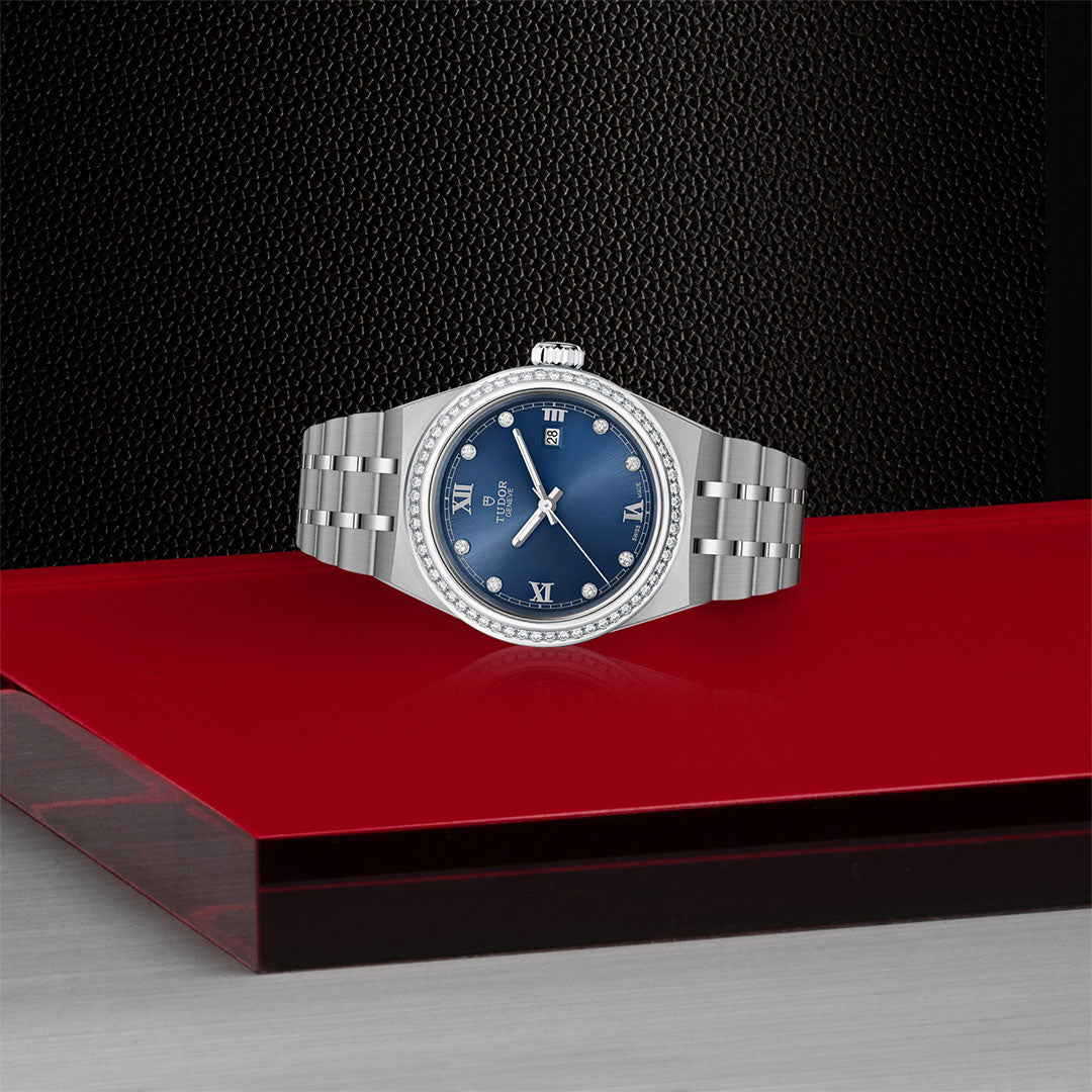 The TUDOR Royal by TUDOR, featuring a blue diamond-set dial with Roman numerals and a silver metal link band, gleams on a glossy red surface against a dark textured background.