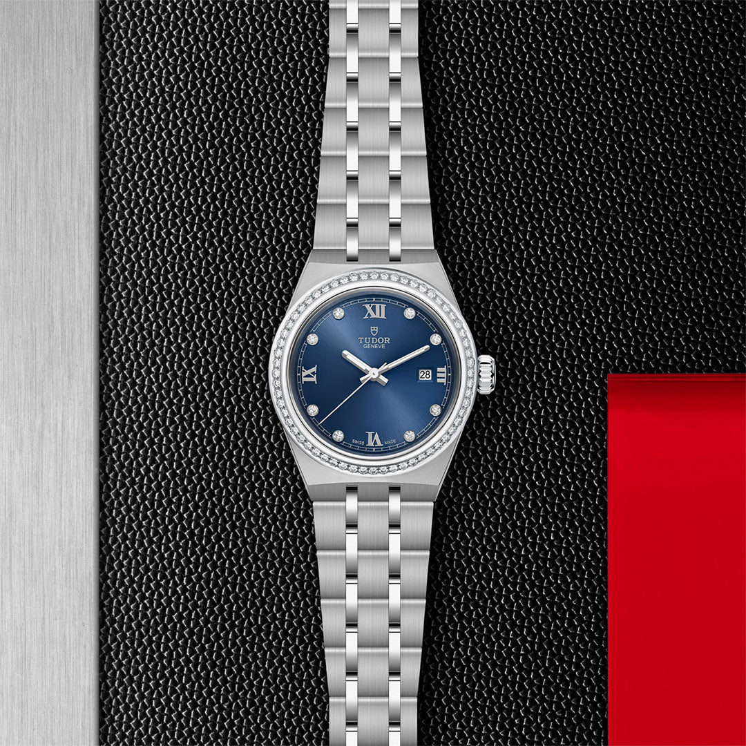 The TUDOR Royal by TUDOR features a stainless steel band, a blue diamond-set dial with silver Roman numerals, diamond hour markers, and a date window, showcased on a textured black and metallic background with a red accent.