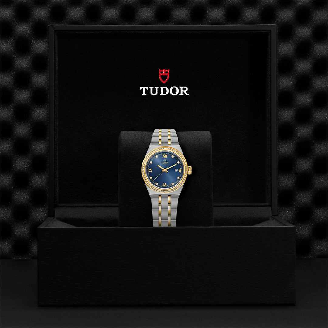 A TUDOR Royal wristwatch with a blue diamond-set dial and gold accents is shown upright in a black TUDOR box featuring the red shield logo and white lettering on the inner lid.