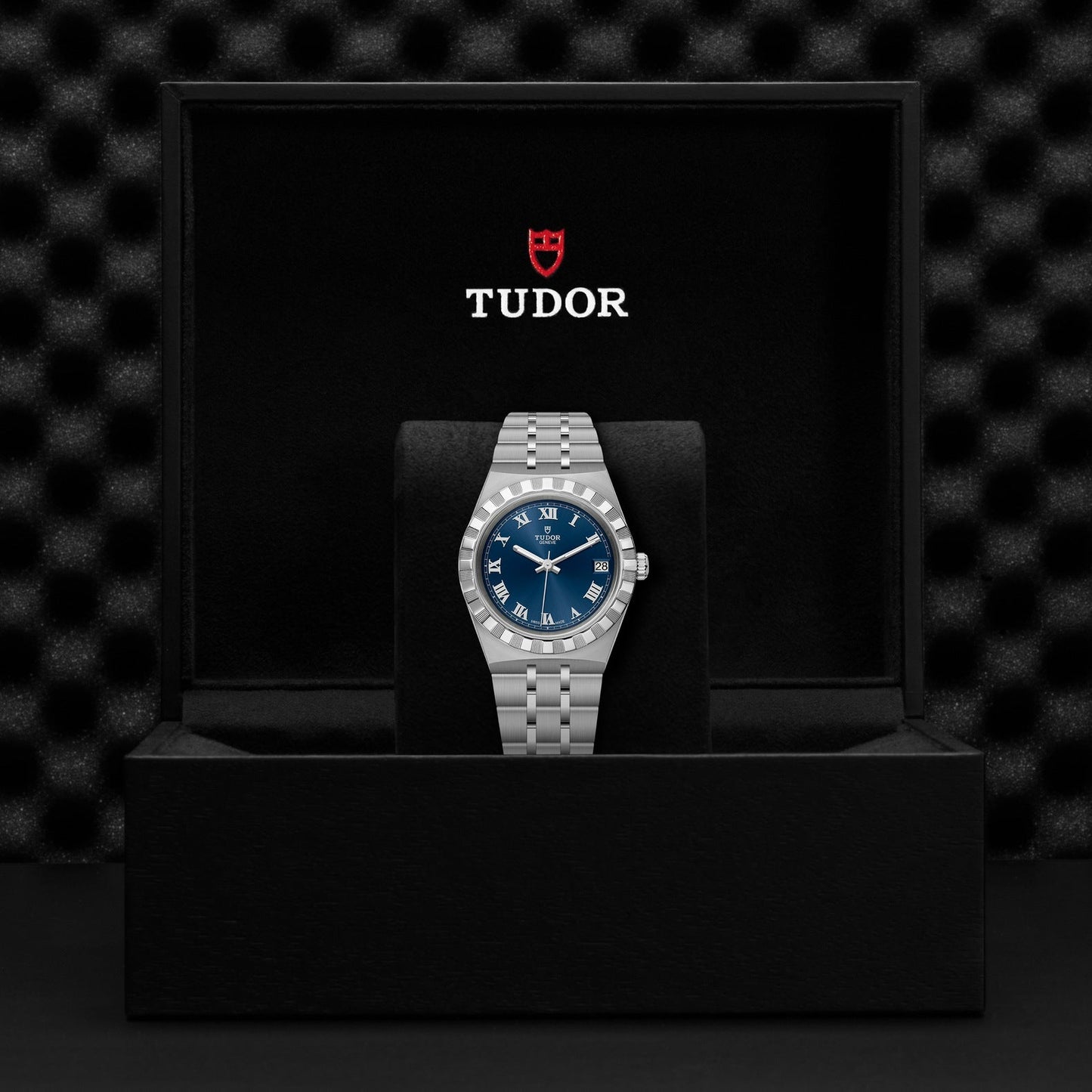 The TUDOR Royal wristwatch, featuring a blue dial and a stainless steel and gold strap, is presented upright in an open black box. The box displays the Tudor logo above the timepiece, set against a textured black background, highlighting its automatic sport-chic appeal.