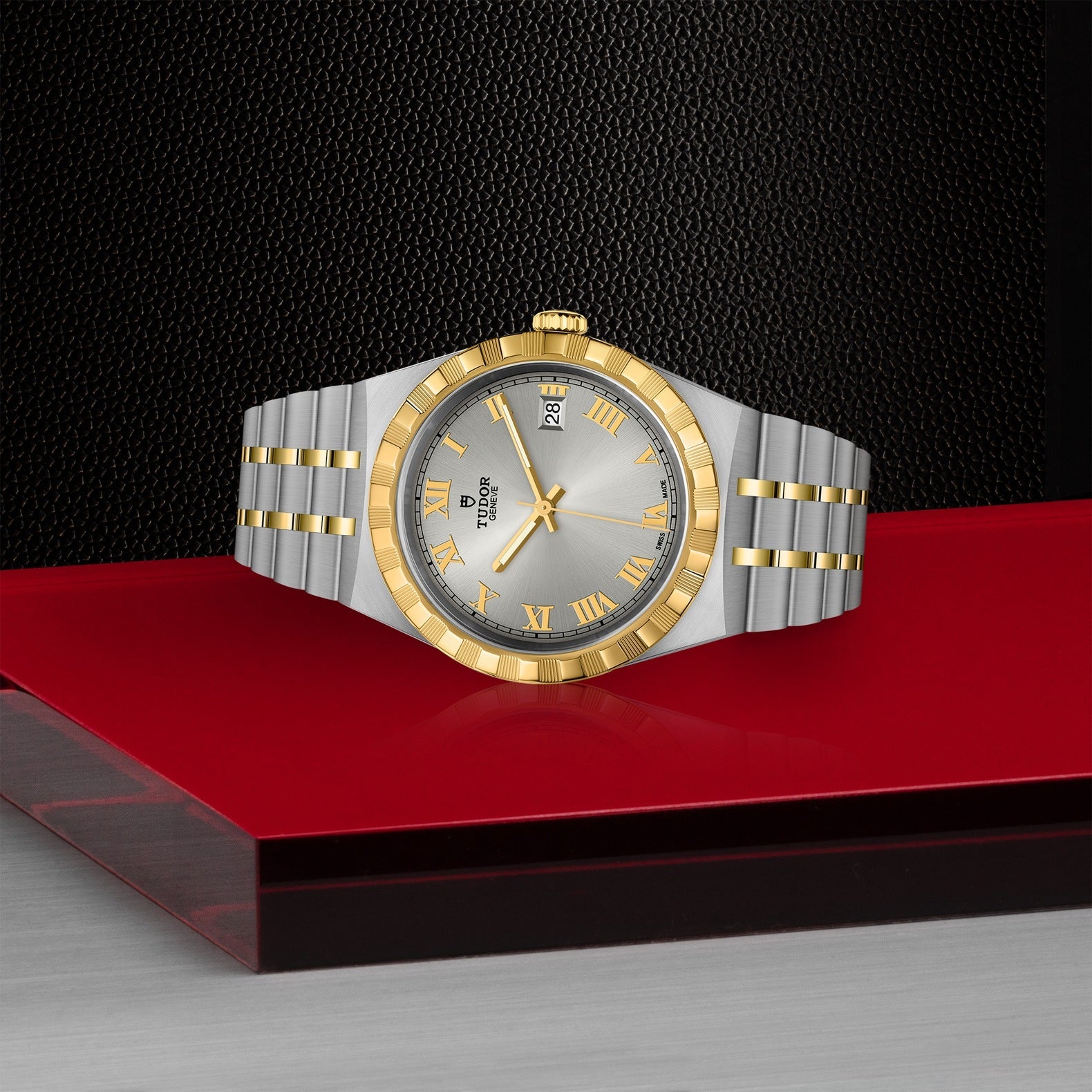 The TUDOR Royal watch elegantly merges stainless steel and gold in its luxurious bracelet, showcasing a gold bezel and a silver dial adorned with gold Roman numerals and the TUDOR logo. It is artfully presented on a red surface with a dark, textured background.