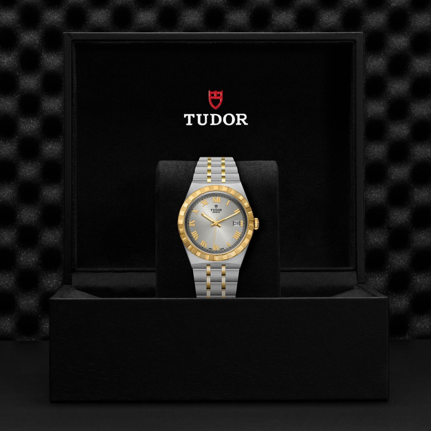 A TUDOR Royal watch is elegantly presented in an open black box, featuring a stainless steel and gold bracelet alongside a gold and silver face. The interior prominently displays the TUDOR logo in white and red, embodying the essence of automatic sport-chic timepieces.