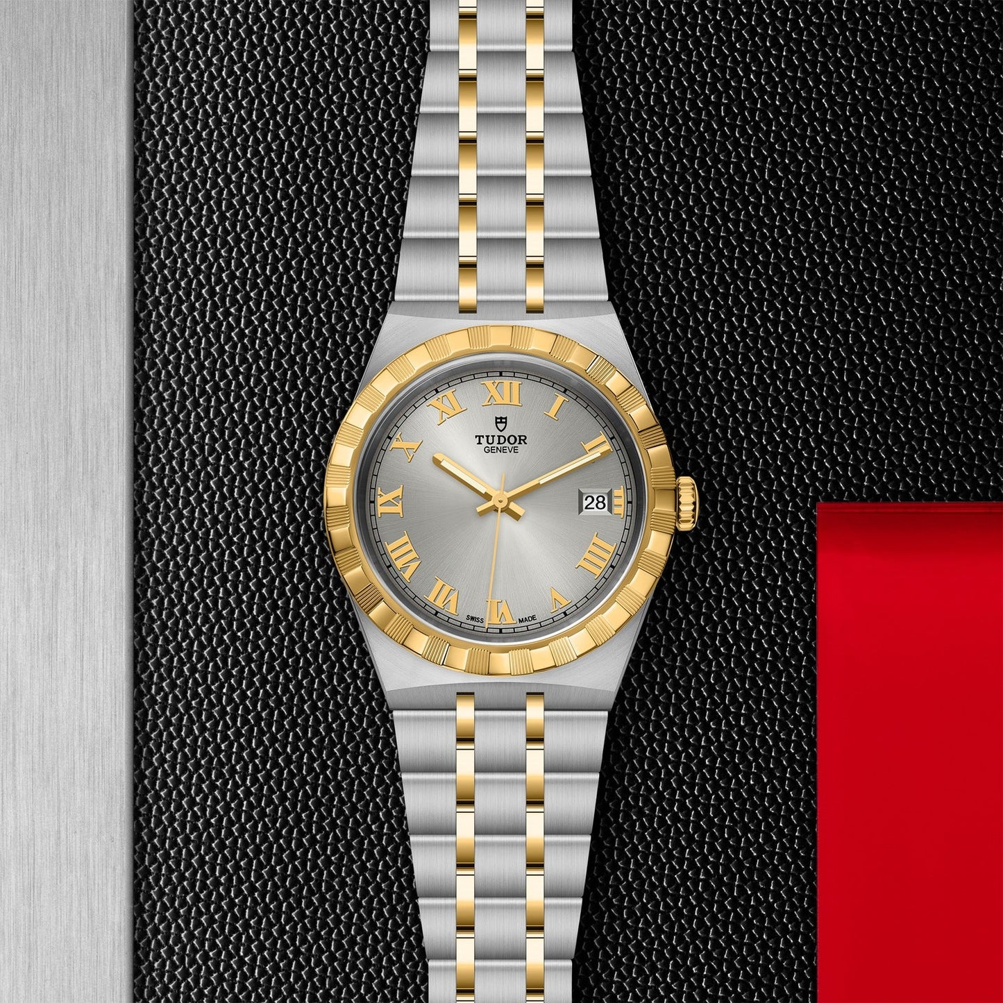 The TUDOR Royal watch by TUDOR showcases a sophisticated silver and gold design, highlighted by a round dial bordered in gold and featuring a date display. Its sport-chic style is further enhanced by the stainless steel and gold link bracelet, set against a textured black background with a metallic strip on the left side and accented with a red square on the right.