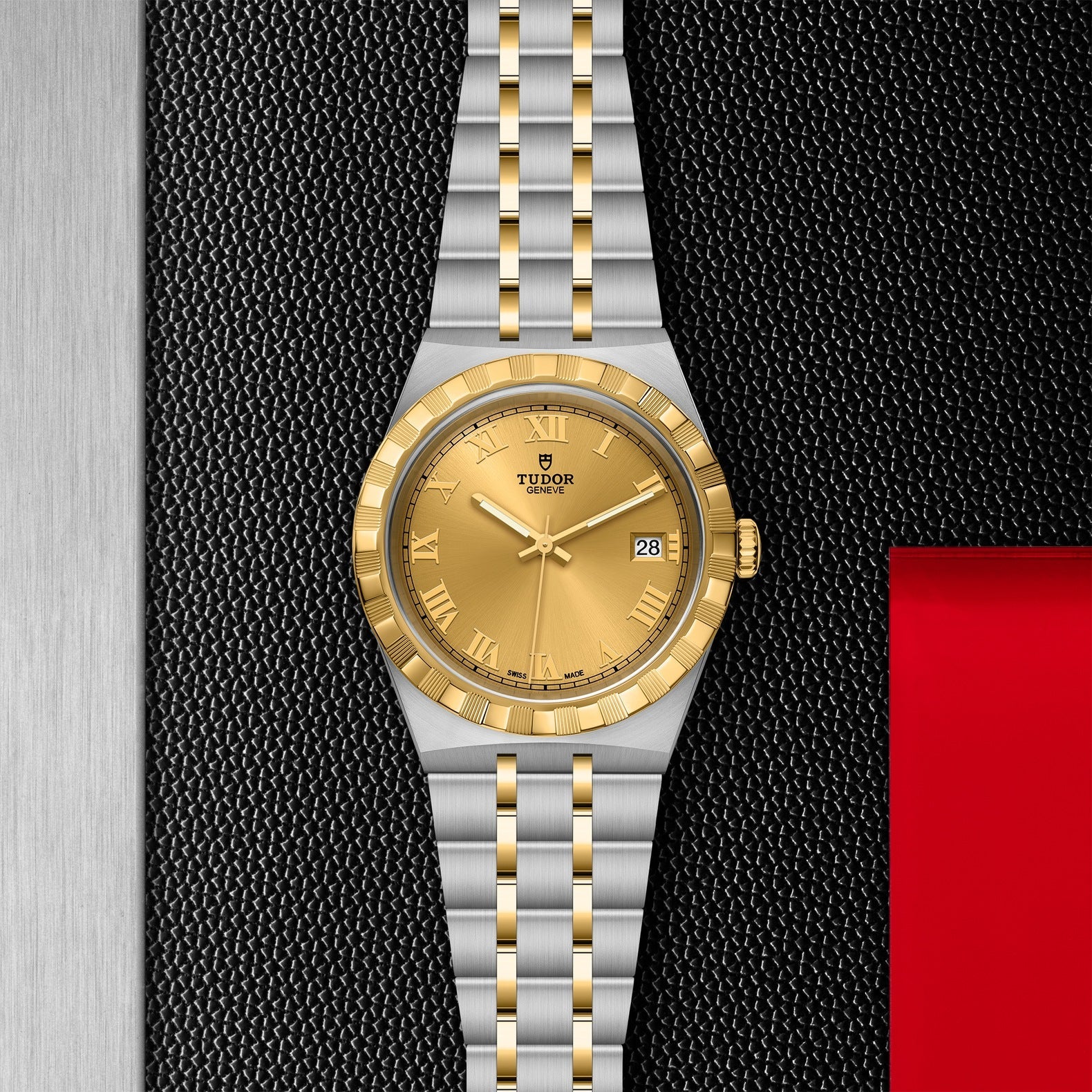 A TUDOR Royal watch from the TUDOR brand features a combination of stainless steel and gold, with a gold dial adorned with Roman numerals and a date function. It is placed on a textured black backdrop, flanked by a shiny red square to the right and a metallic gray edge to the left.