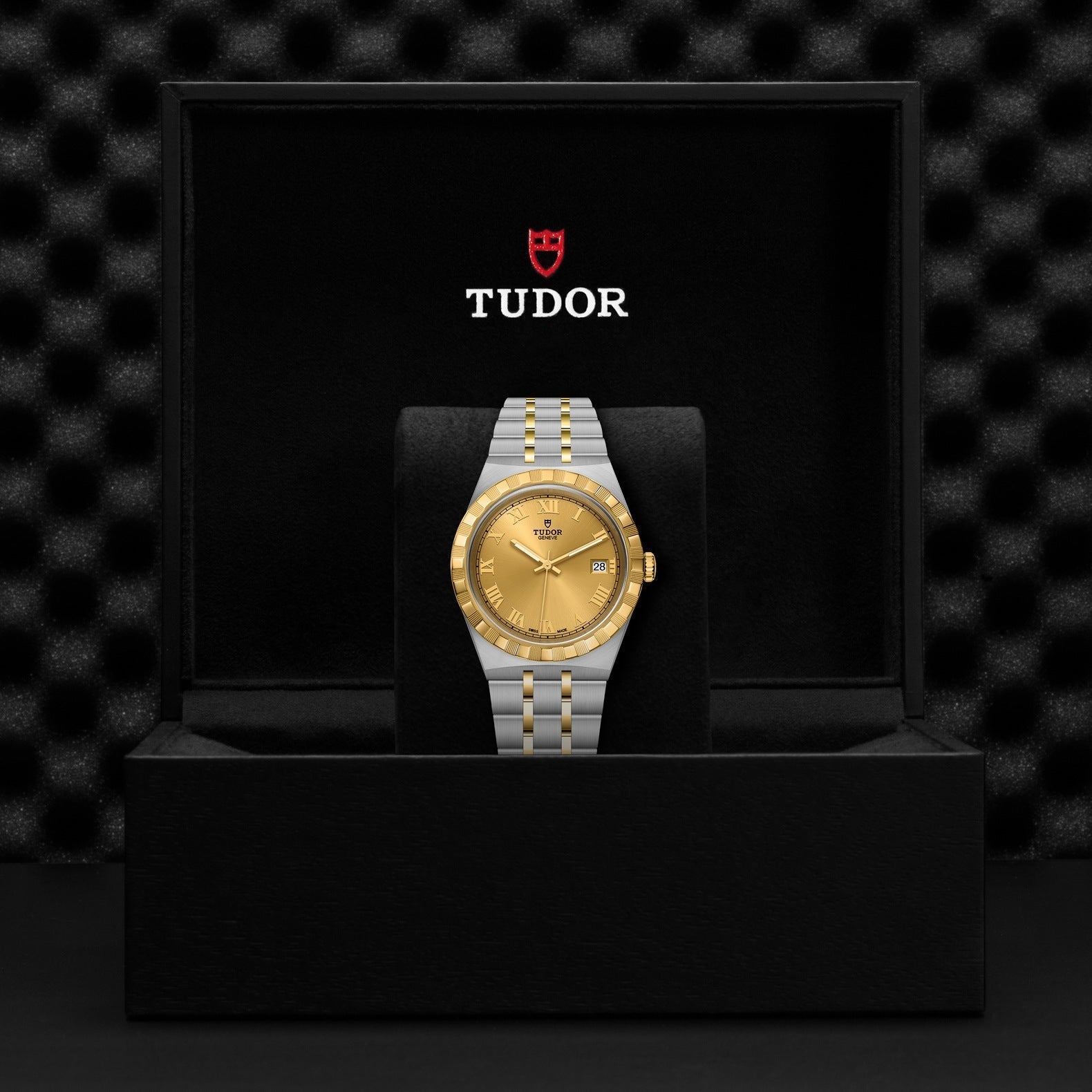 A TUDOR Royal timepiece, featuring a gold dial and a dual-tone stainless steel and gold bracelet, is elegantly presented in an open black box. The interior highlights the TUDOR logo above the watch against a richly textured black backdrop, embodying a blend of elegance and sport-chic design.